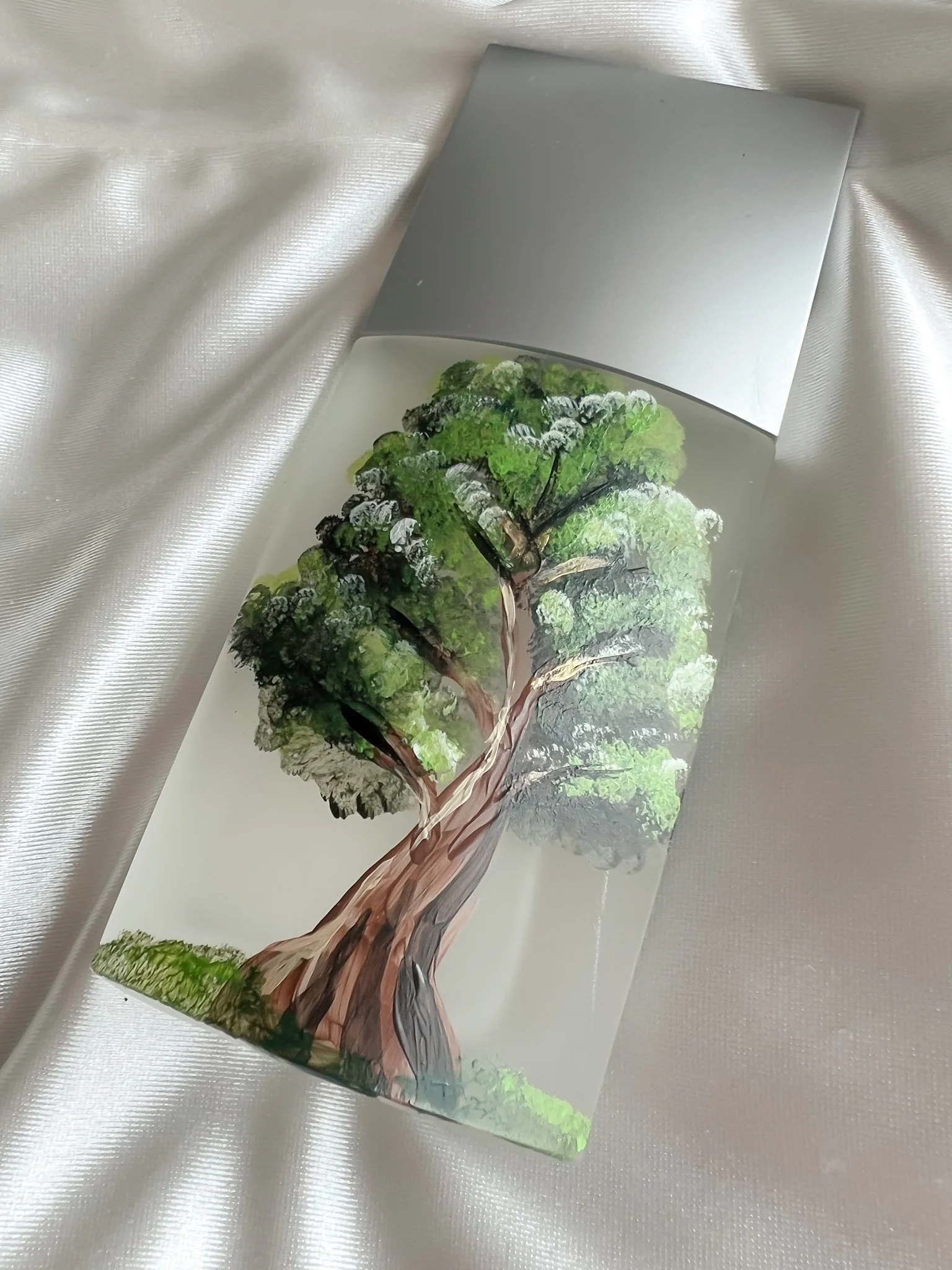 Box of preserved green and brown moss or lichen with a tree painting on the front, placed on a white satin fabric.