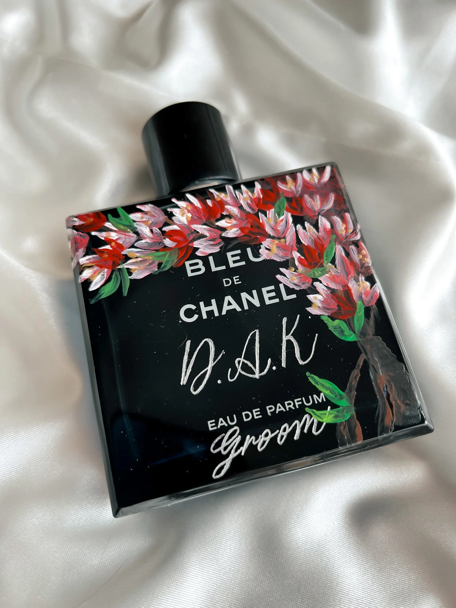 A black Chanel perfume bottle decorated with pink, red, and green painted floral designs, placed on a white satin fabric.