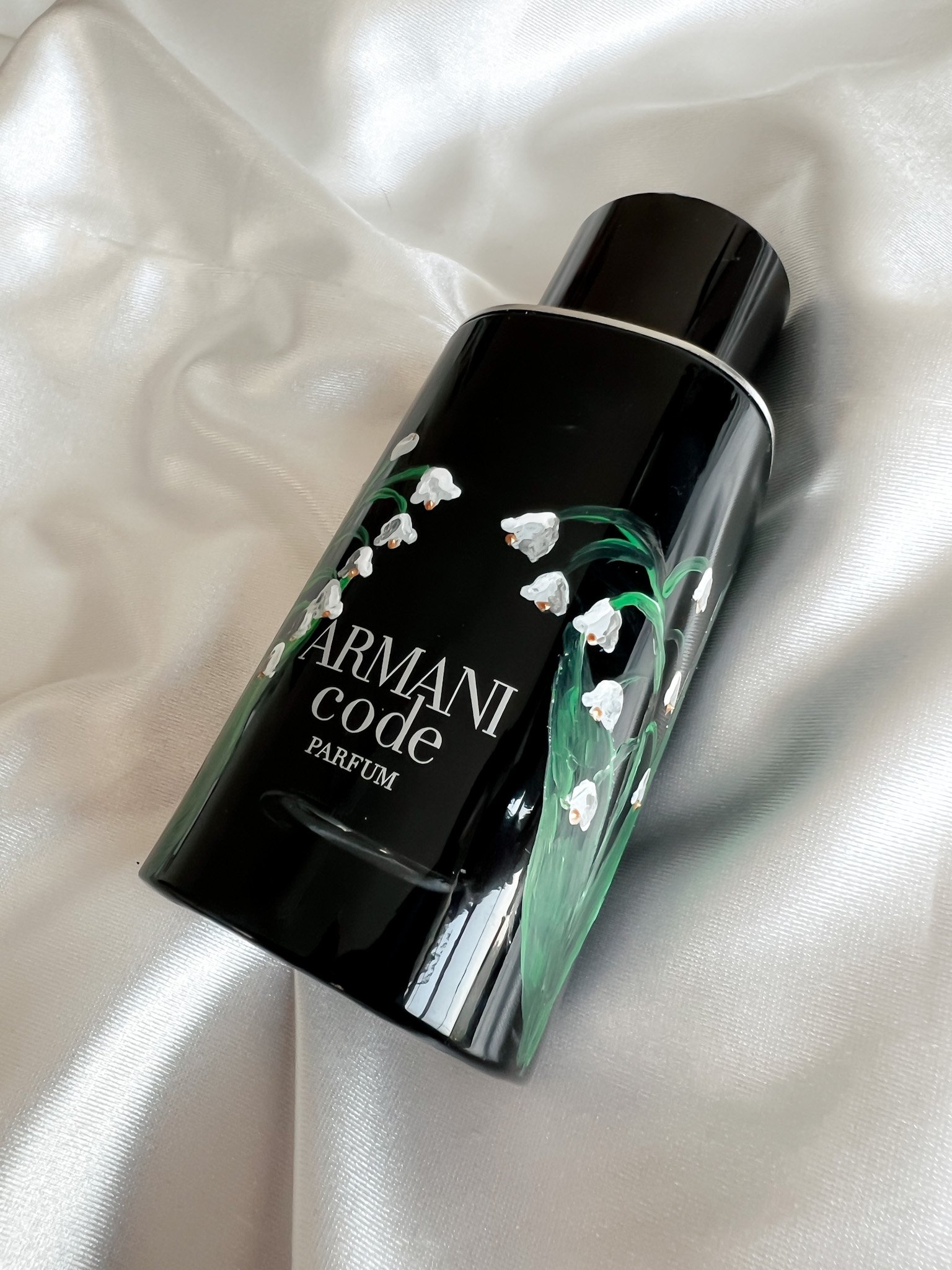 A black bottle of Armani Code perfume with lily of the valley flowers painted on it, lying on cream satin fabric.