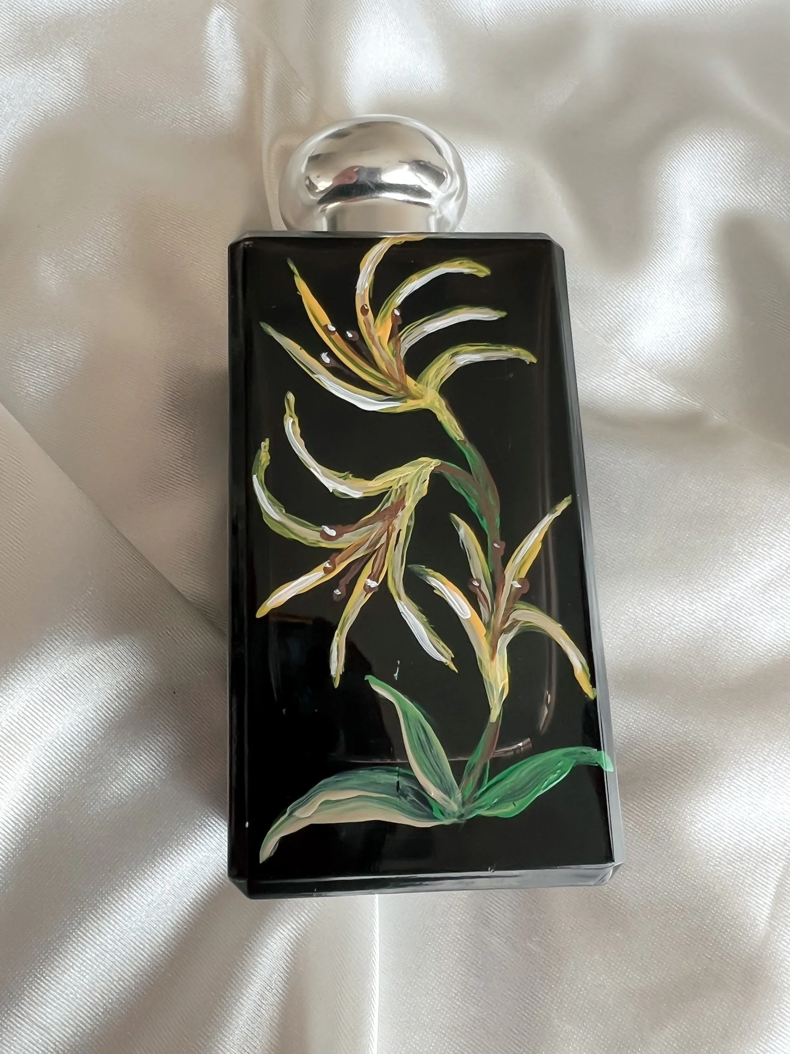 A black perfume bottle with painted floral design and a clear glass cap, placed on a shiny white fabric surface.