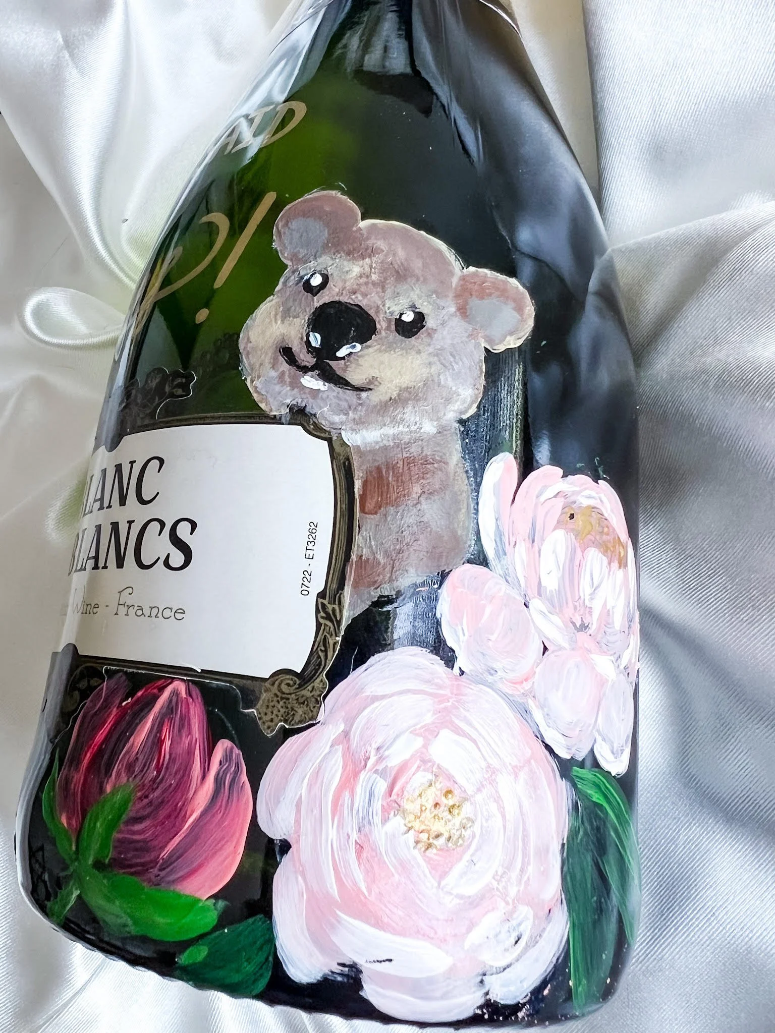 Decorative wine bottle with hand-painted illustrations of a smiling bear and pink flowers, wrapped in a shiny bag on a satin background.