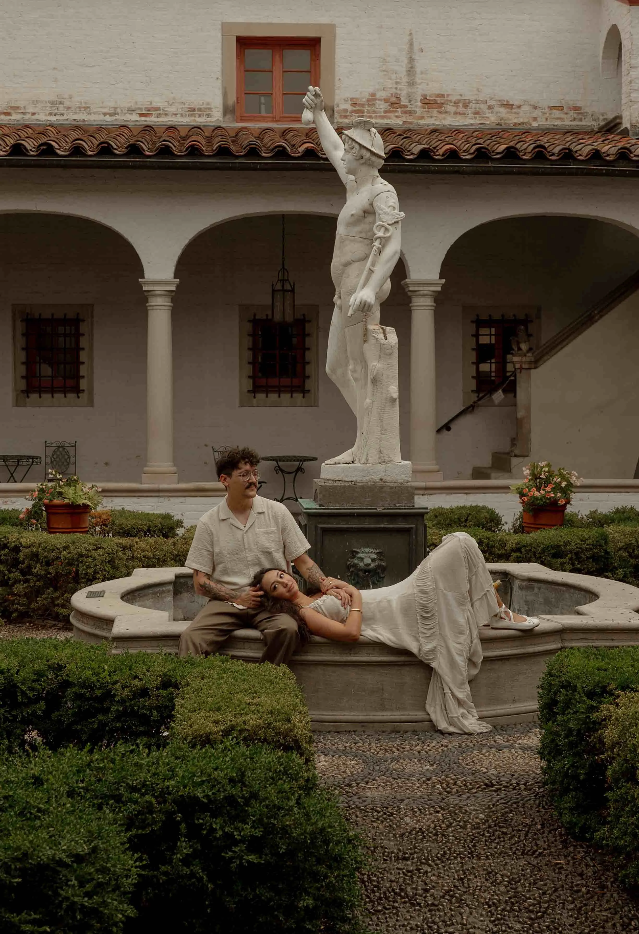 A man with a mustache and glasses sitting on the edge of a fountain holding a woman in a white dress lying back in his lap. Behind them is a stone statue of a naked man with a sword, in a courtyard with bushes and flowers.