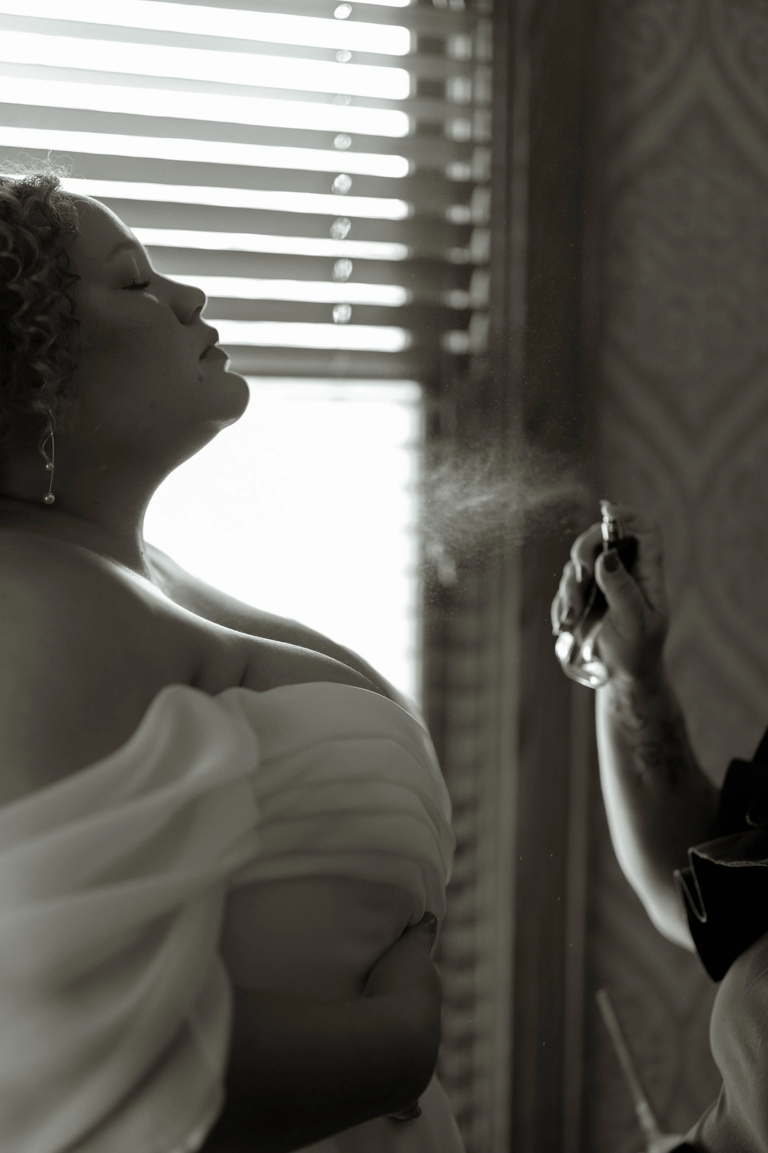A woman with curly hair in profile, wearing earrings and a white off-shoulder wedding gown, spraying perfume in a room with horizontal blinds, in black and white.