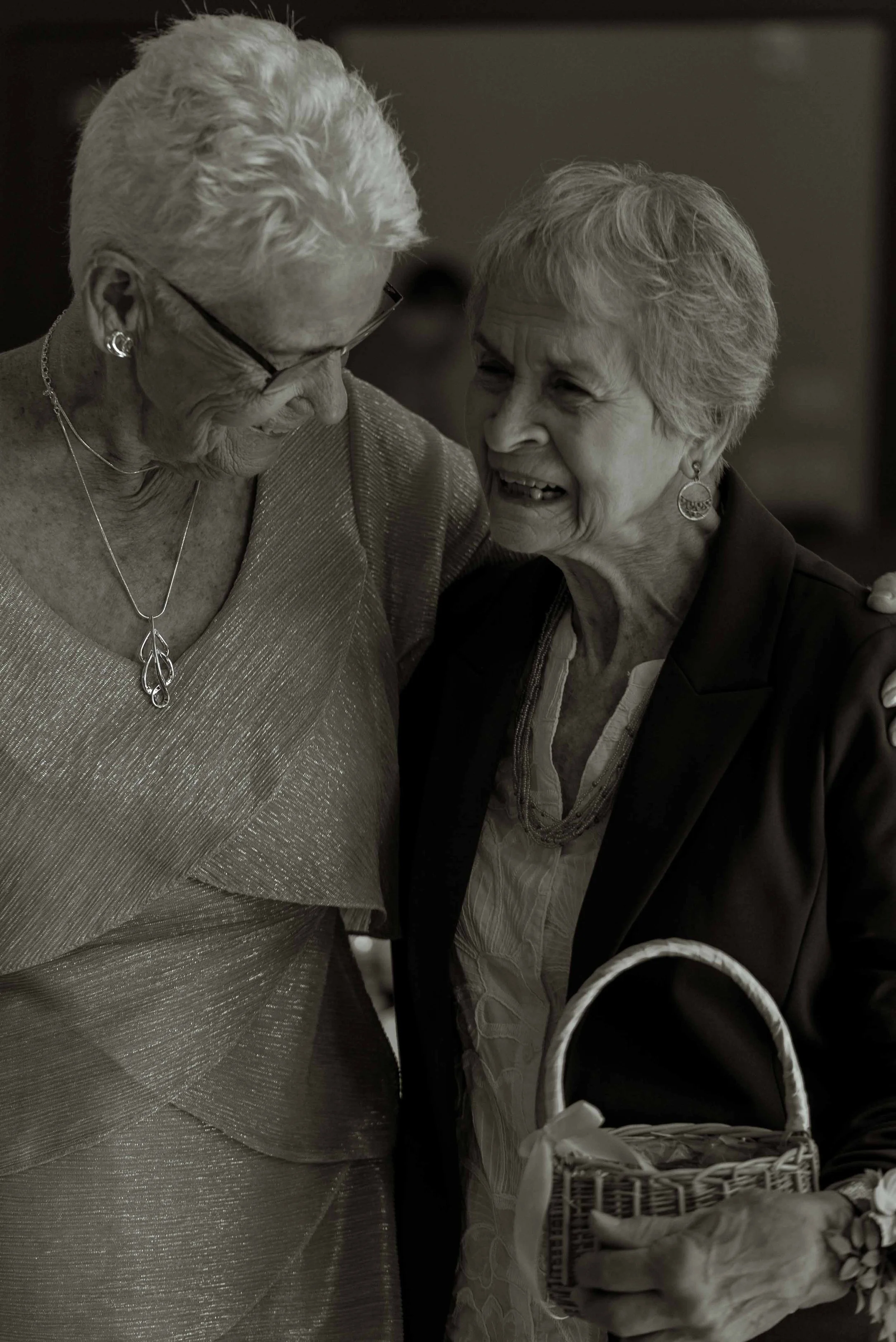 Two elderly women smiling and sharing a moment together, one holding a basket of flowers