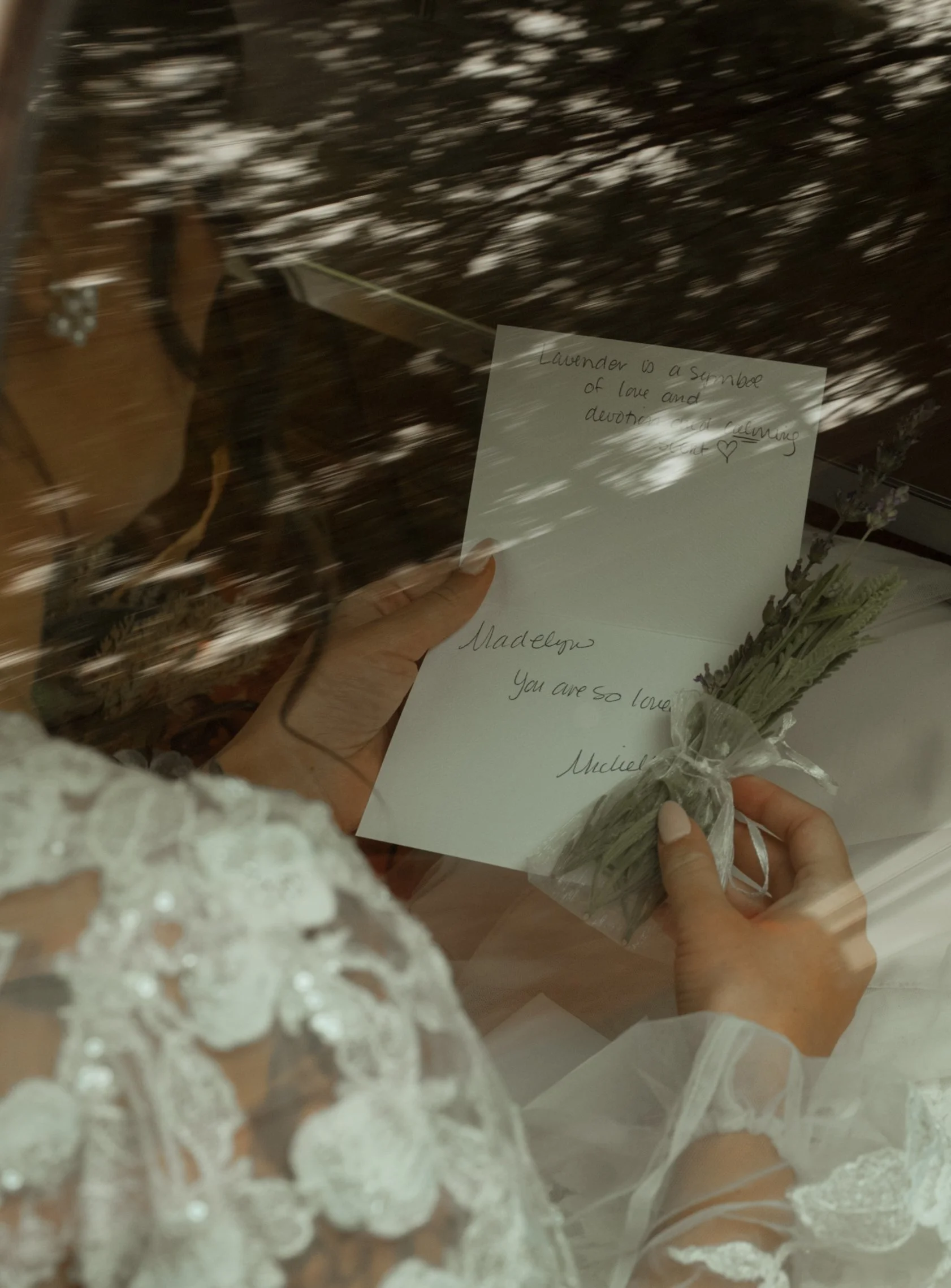 A woman in a lace dress holding a handwritten note and a small bouquet of lavender behind a glass window, with reflections of trees outside.