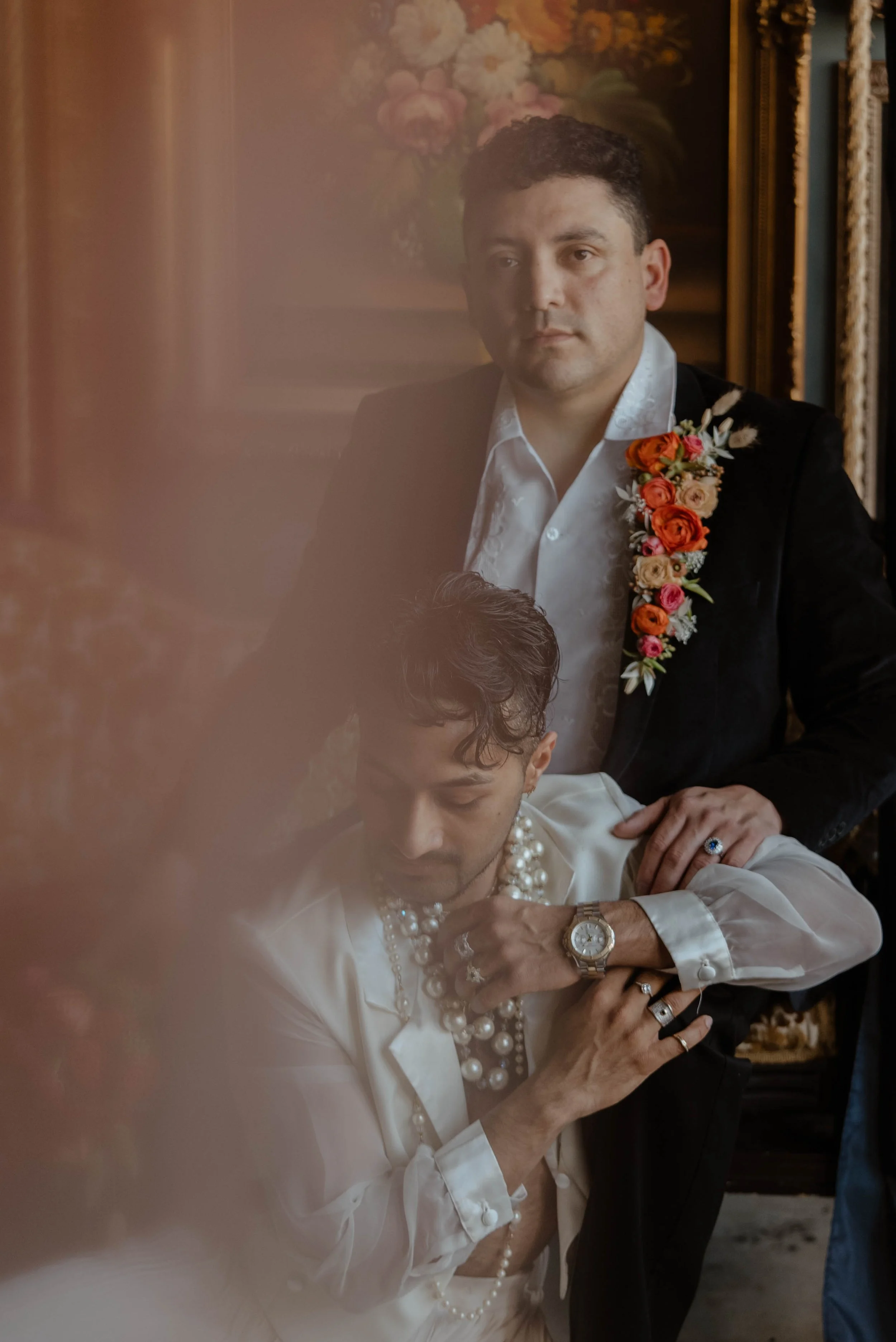 Two grooms in formal wedding attire, one wearing a black jacket with a floral boutonniere, and the other in a light-colored shirt with pearls and jewelry, engaged in a moment of reflection in an ornate room with floral artwork background.