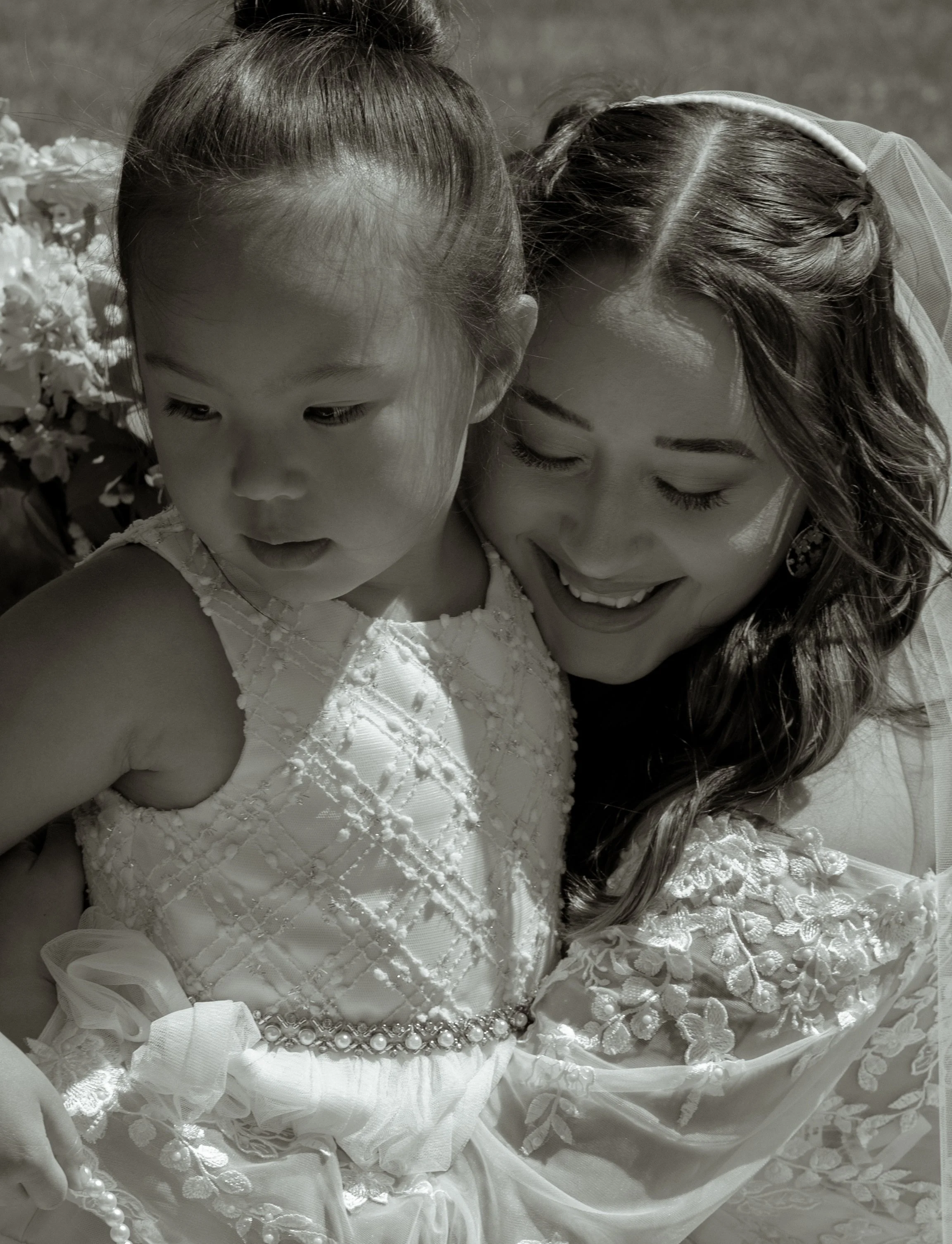 A young girl and a woman sharing a joyful moment, with the woman smiling and the girl looking down, both dressed in fancy clothes.