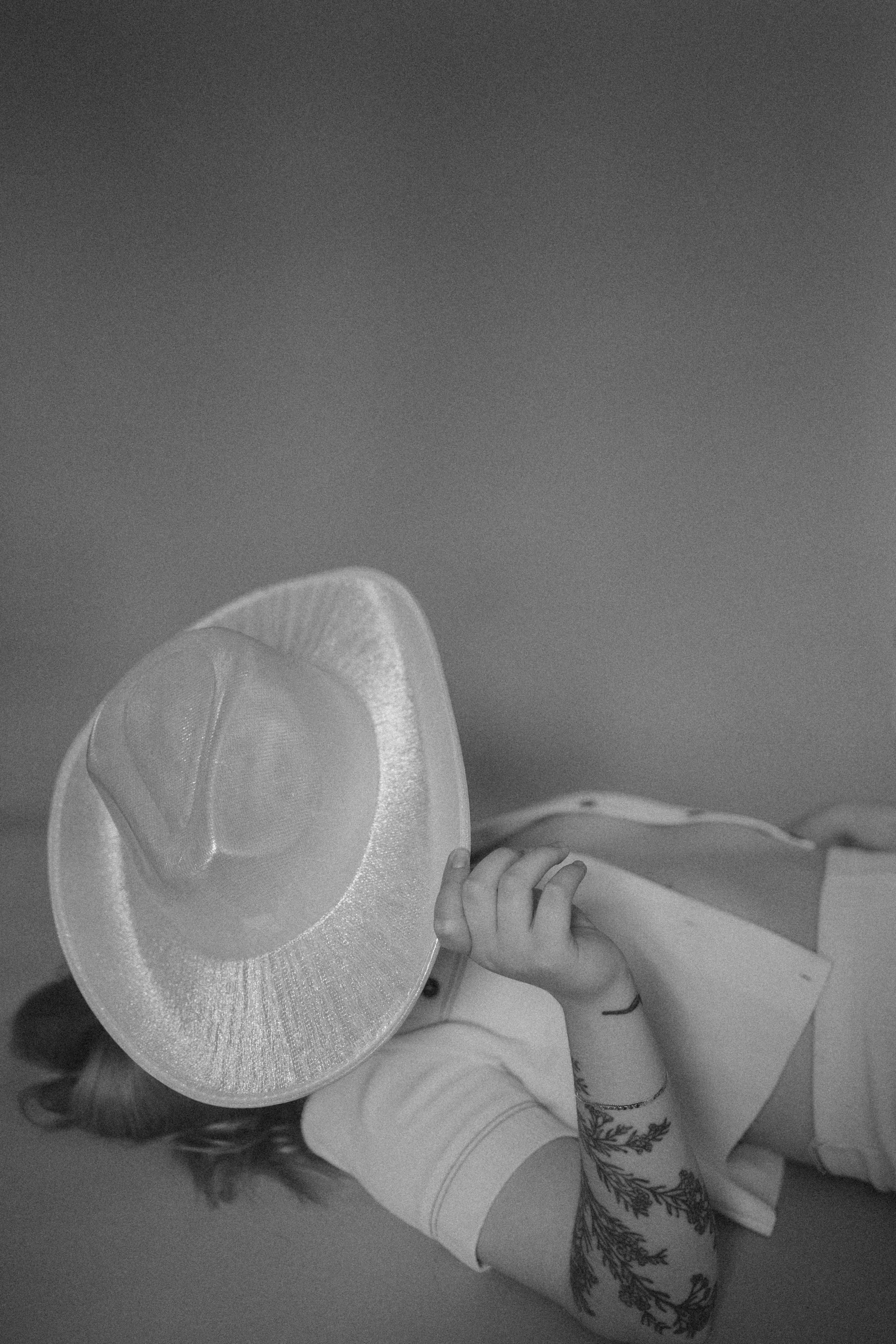 A woman wearing a large cowboy hat covering her face, lying down on her side, with her hand resting on her hat, showing tattoos on her forearm.