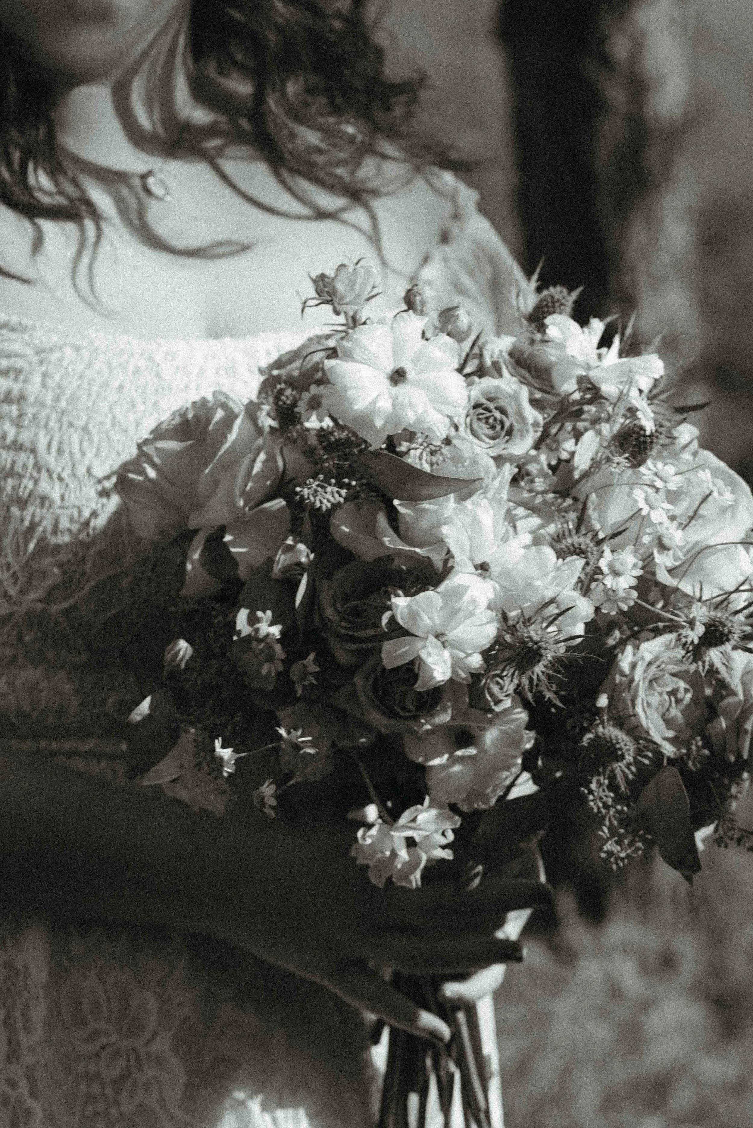 A close-up of a wedding bouquet with various flowers including roses, small white flowers, and greenery, held by a person with dark, curly hair.