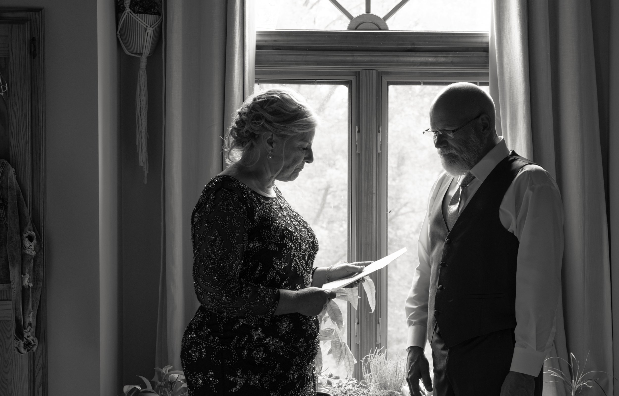 A woman and a man stand facing each other in front of a window, with the woman reading from a piece of paper.