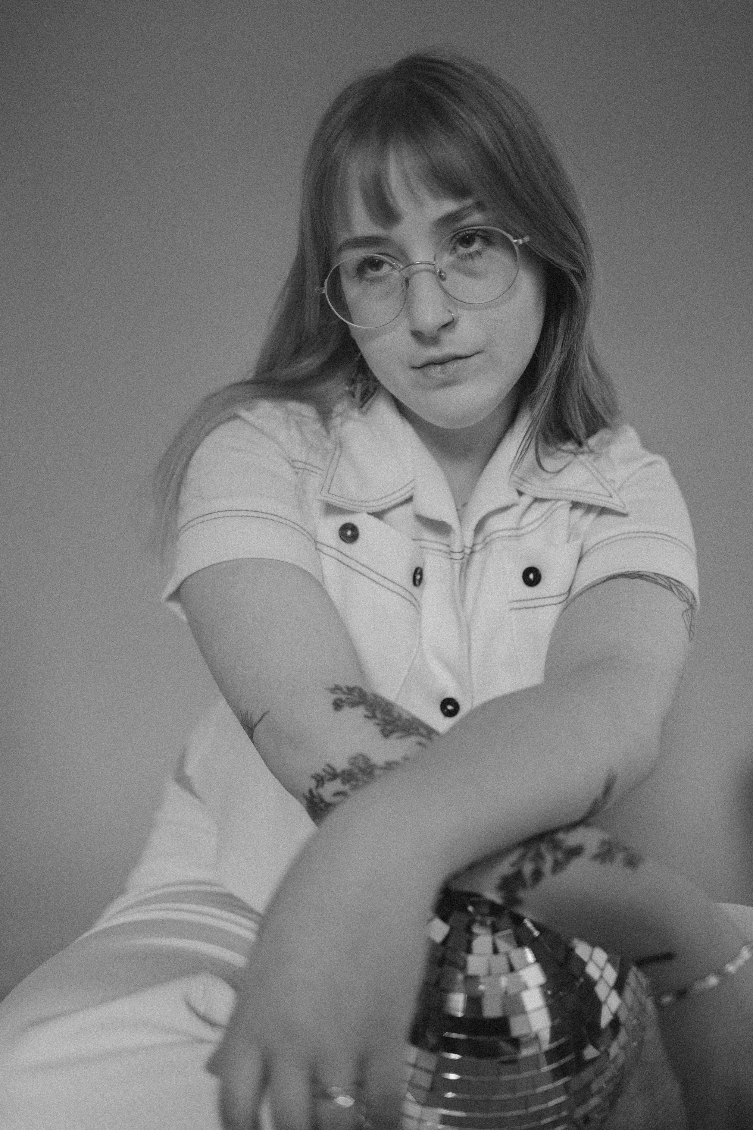 Black and white photo of a young woman with glasses and tattoos reaching towards the camera, sitting with her arm resting on a disco ball.