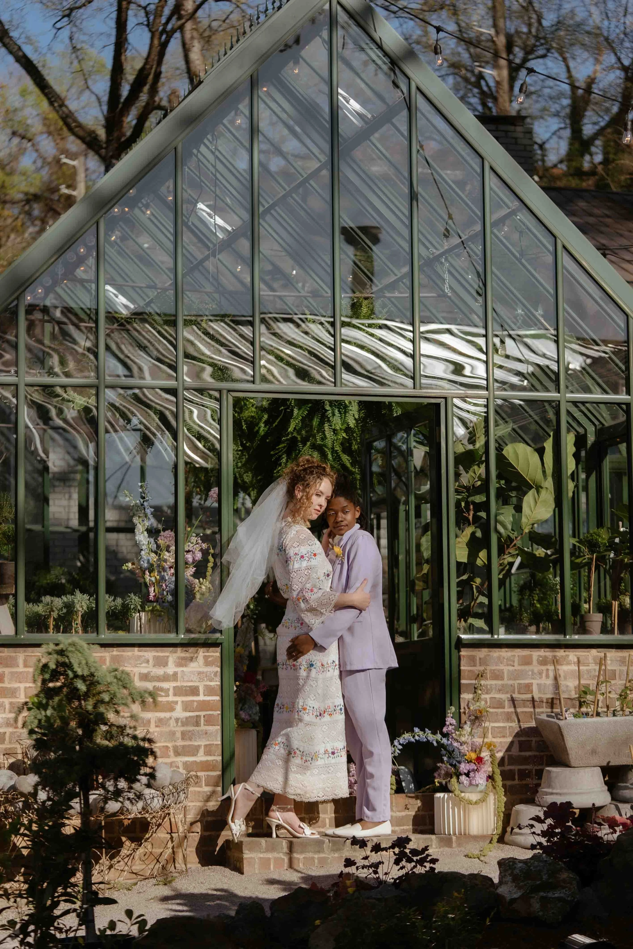 Two women, one in a white lace wedding dress and veil, and the other in a pastel purple suit, pose together outside a glass greenhouse with plants and flowers.