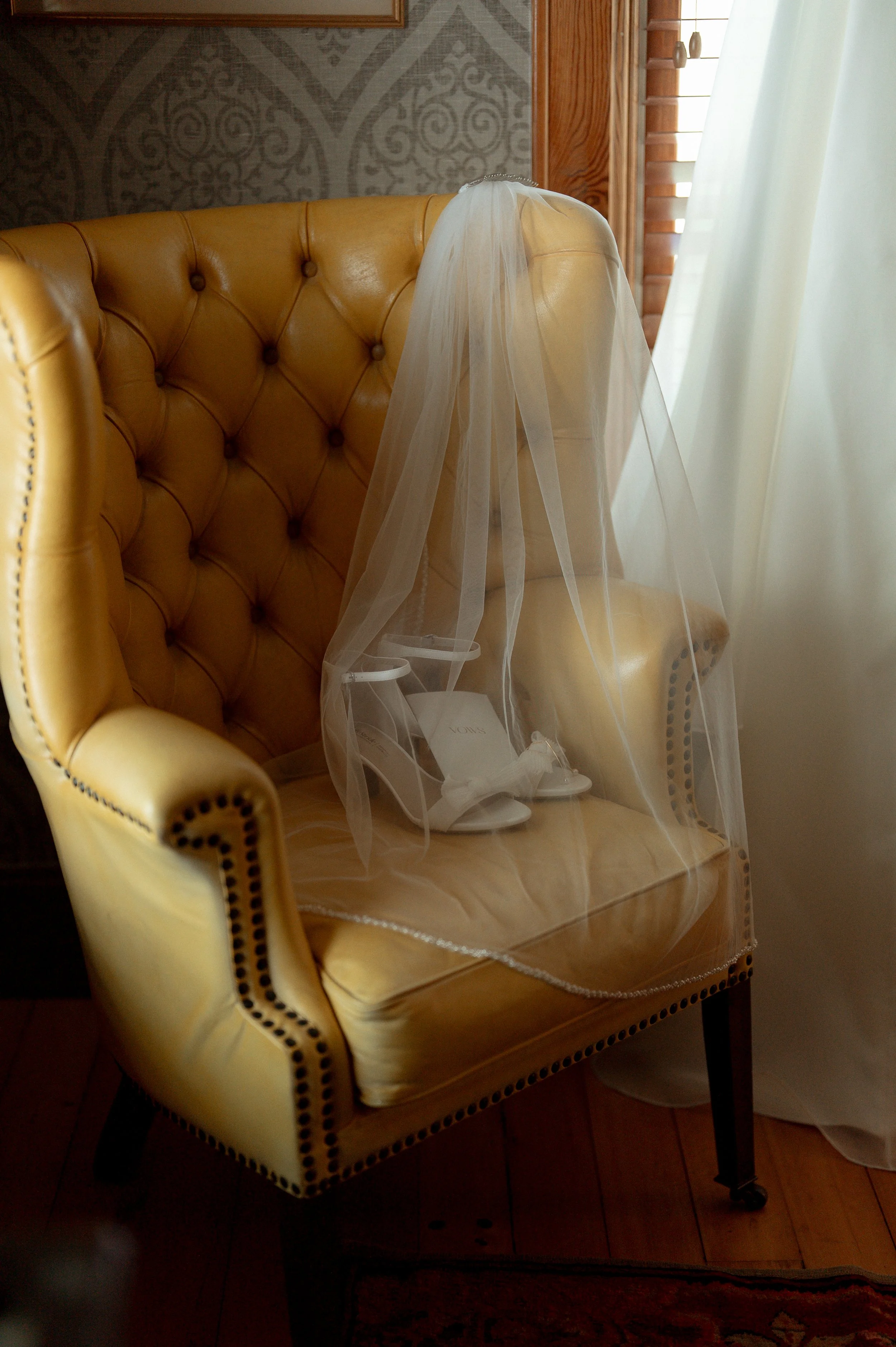 A yellow upholstered armchair with black nailhead trim and a sheer white veil draped over the backrest, placed near a window with white curtains, in a room with patterned wallpaper and wooden accents.
