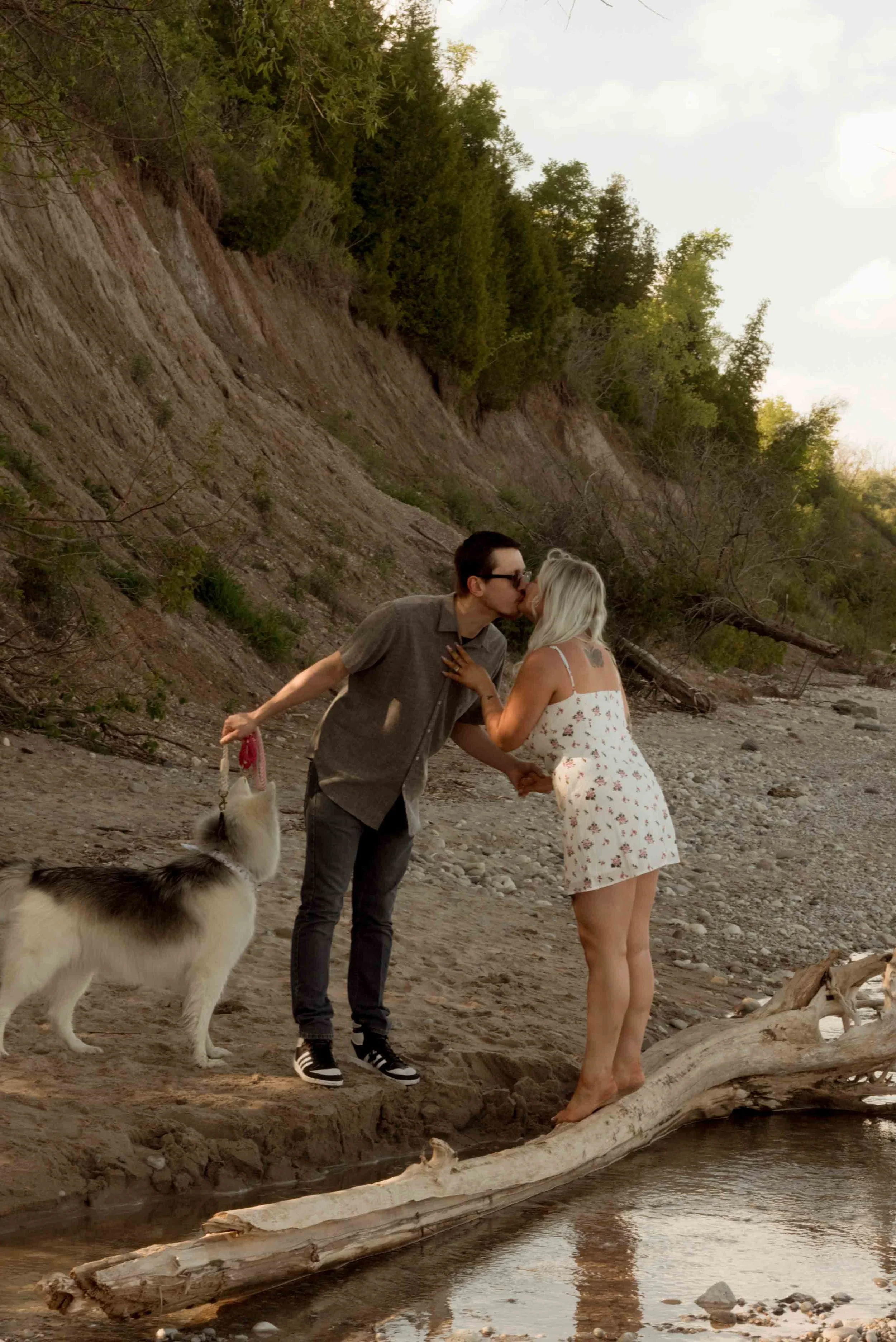 A couple kissing on a riverbank while holding hands, with a dog on a leash nearby, and a wooded hillside in the background.
