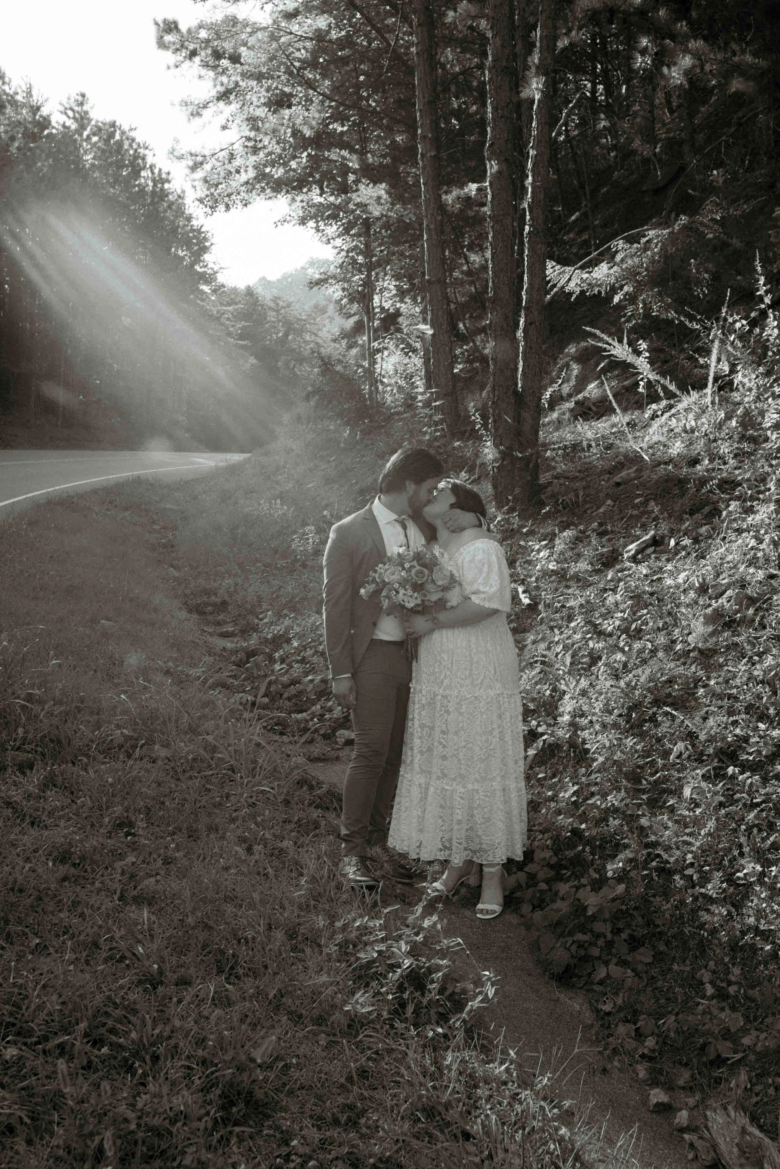 A black and white photo of a couple standing close together on a forested roadside, sharing a kiss. The woman holds a bouquet of flowers, and the sunlight filters through trees in the background.