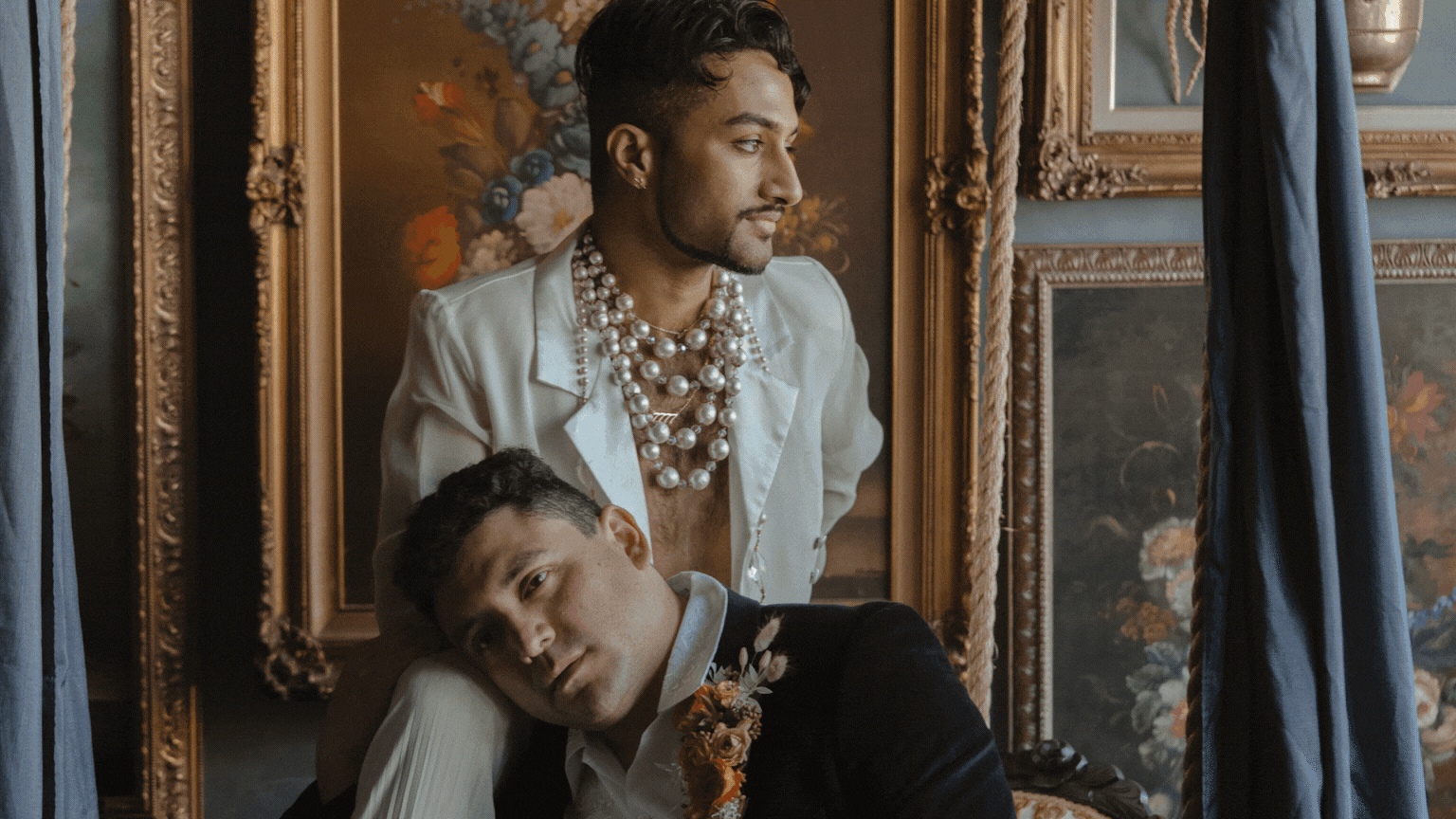 Two men in an art gallery, one leaning on a wall and the other standing, surrounded by framed paintings and blue curtains.