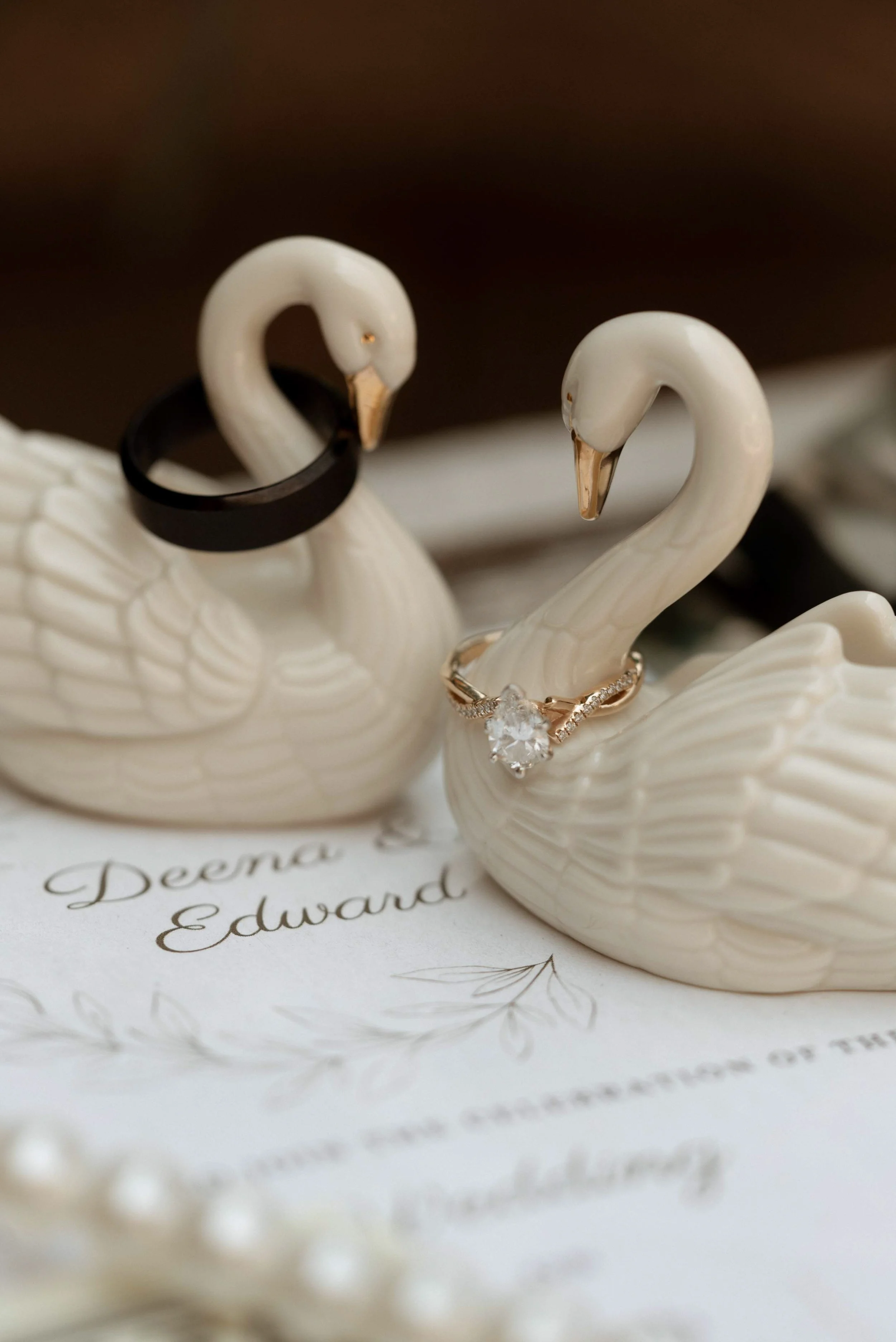 Two ceramic swan figurines, one wearing a black ring and the other an engagement ring with a large central diamond, set on a decorative paper that reads 'Deena' and 'Edward'.