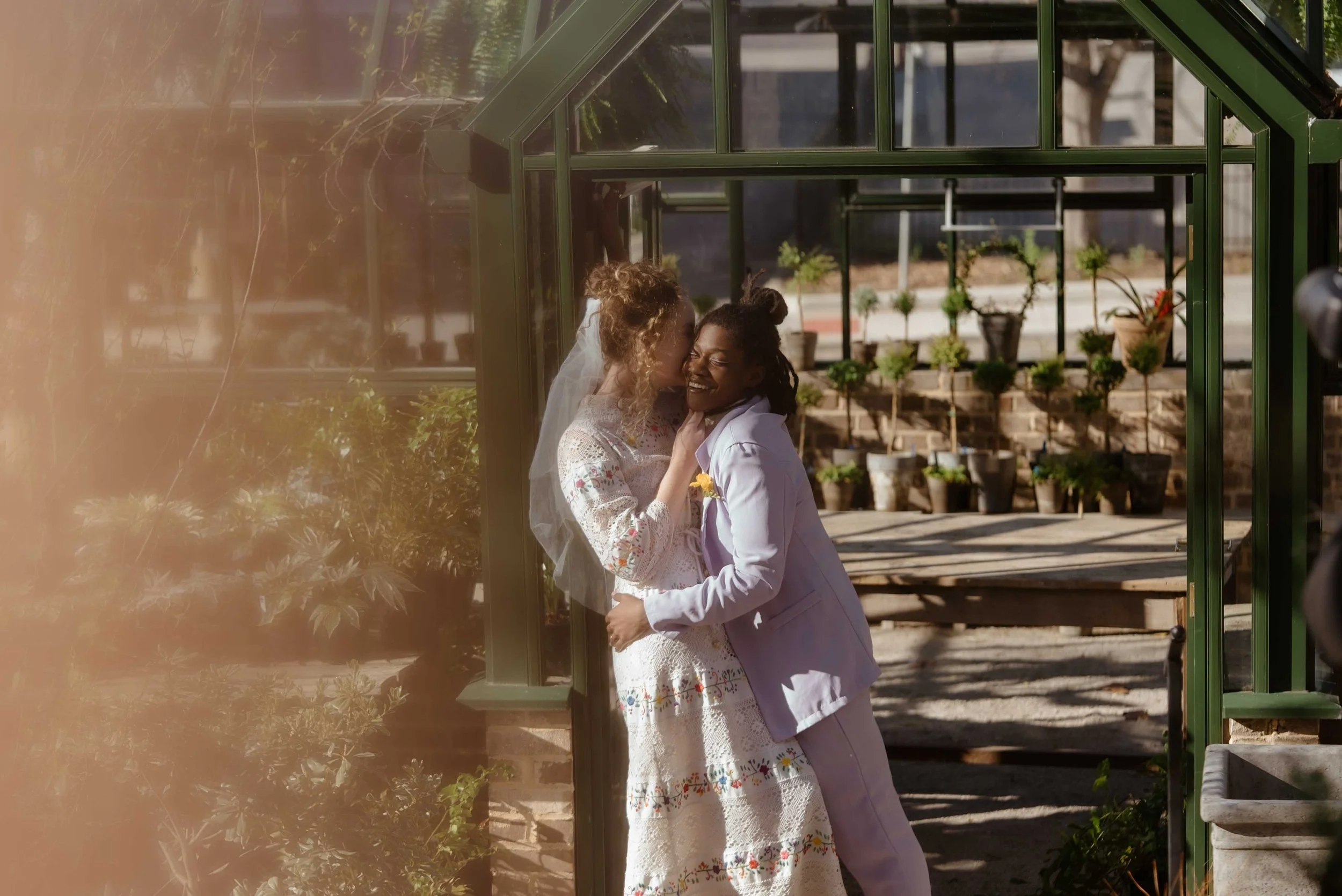 Two women in wedding atitre embrace and share a kiss outside a greenhouse structure with potted plants, bricks, and trees visible in the background.