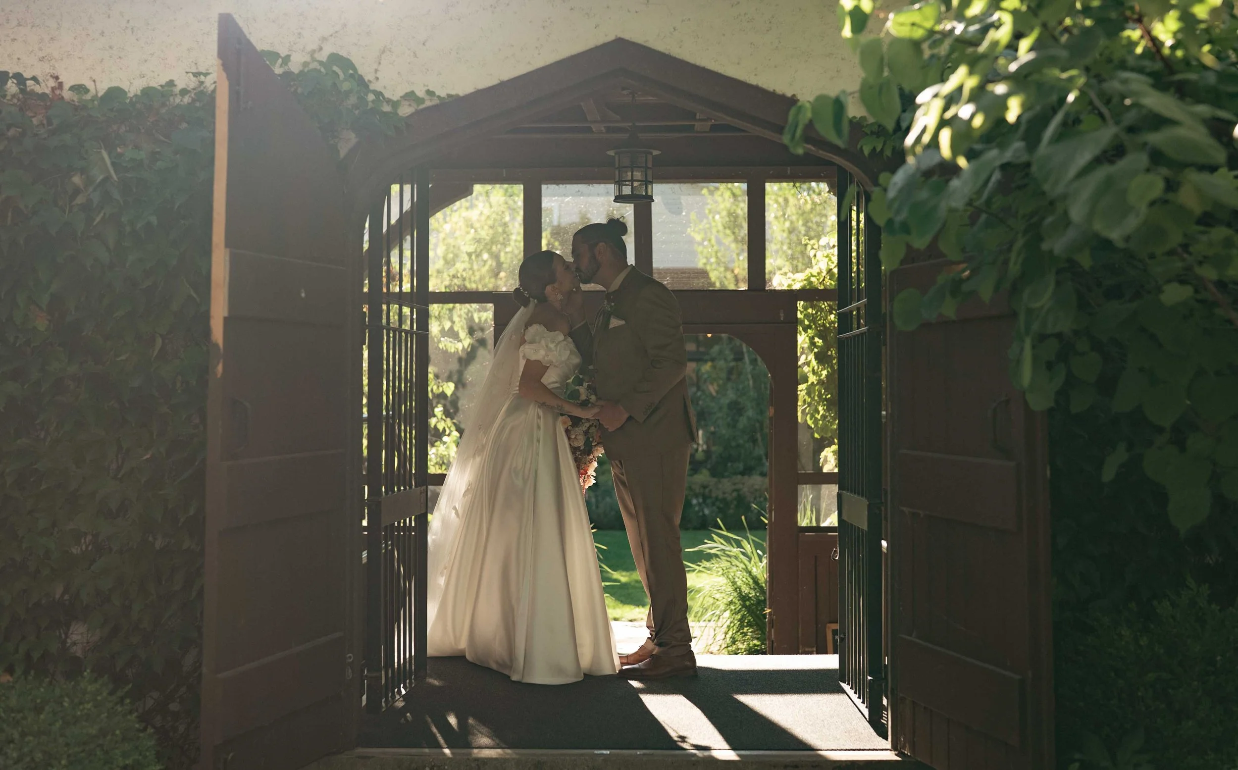 A bride and groom share a kiss under a wooden gazebo, surrounded by greenery and open gates, during a wedding ceremony during golden hour.