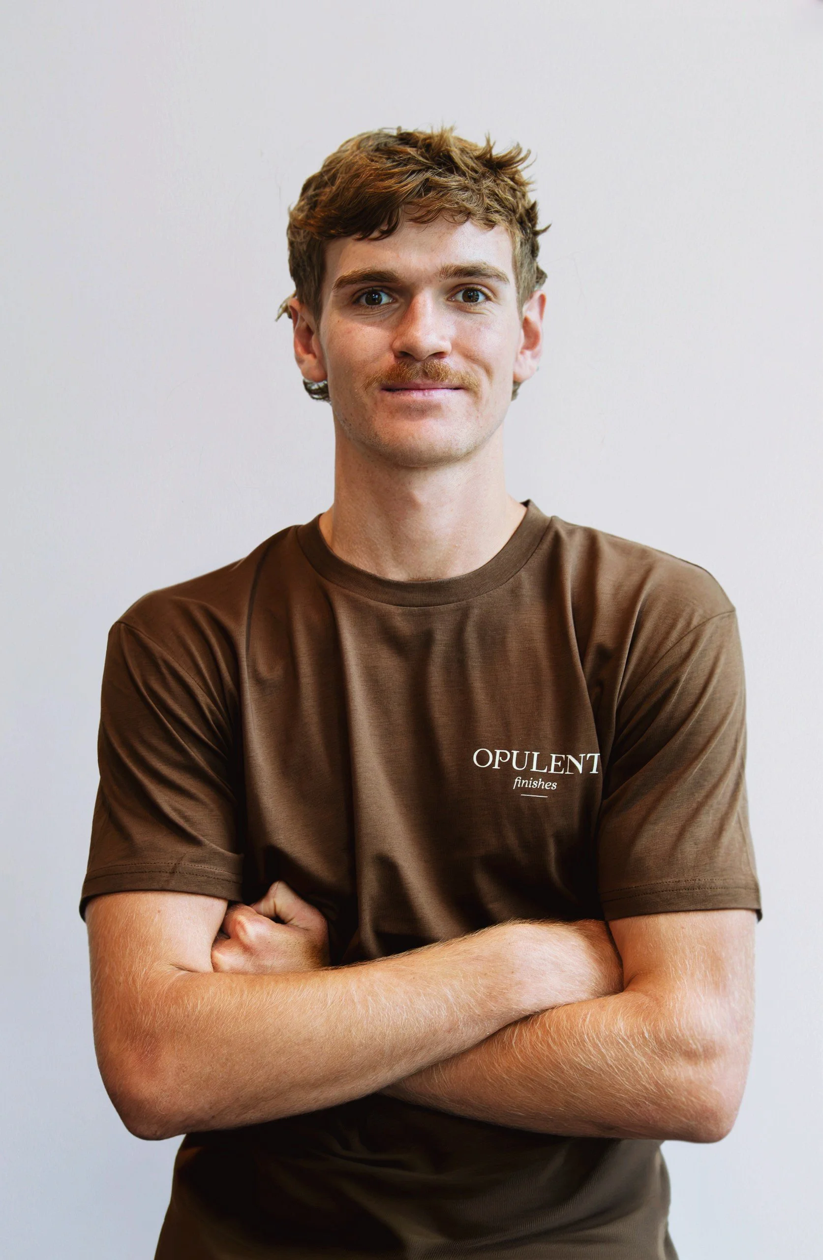 A young man with red hair and a mustache, standing against a plain white wall, wearing a brown "Opulent Finishes" t-shirt with arms crossed.