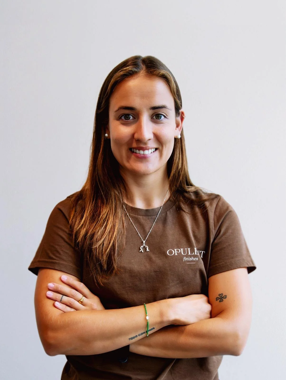 A woman with long brown hair, smiling, wearing a brown t-shirt with 'OPULENT finishes' written on it, standing against a plain white wall.