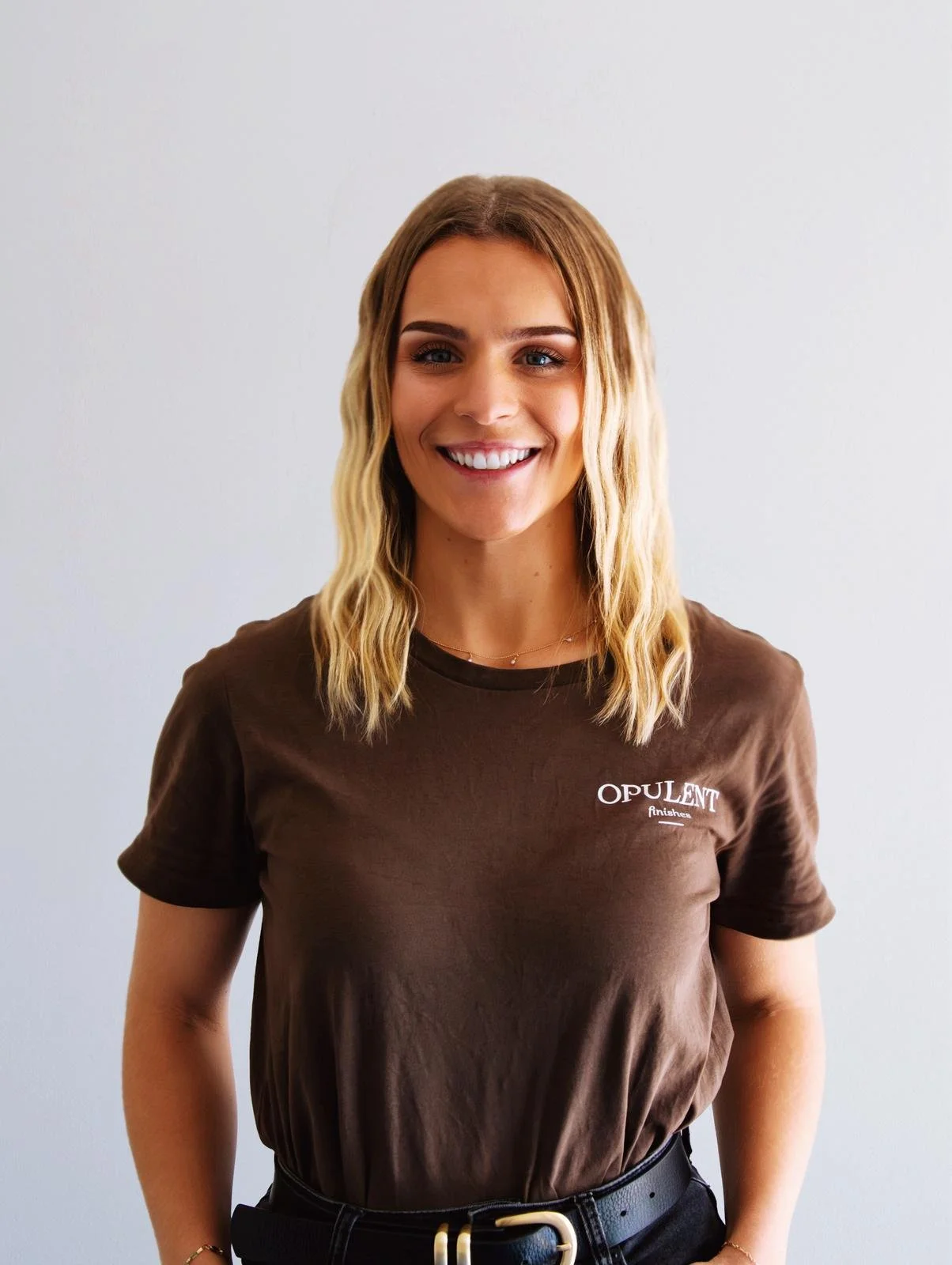 A woman smiling, wearing a brown T-shirt with the logo 'Opulent Finishes' standing against a plain light background.