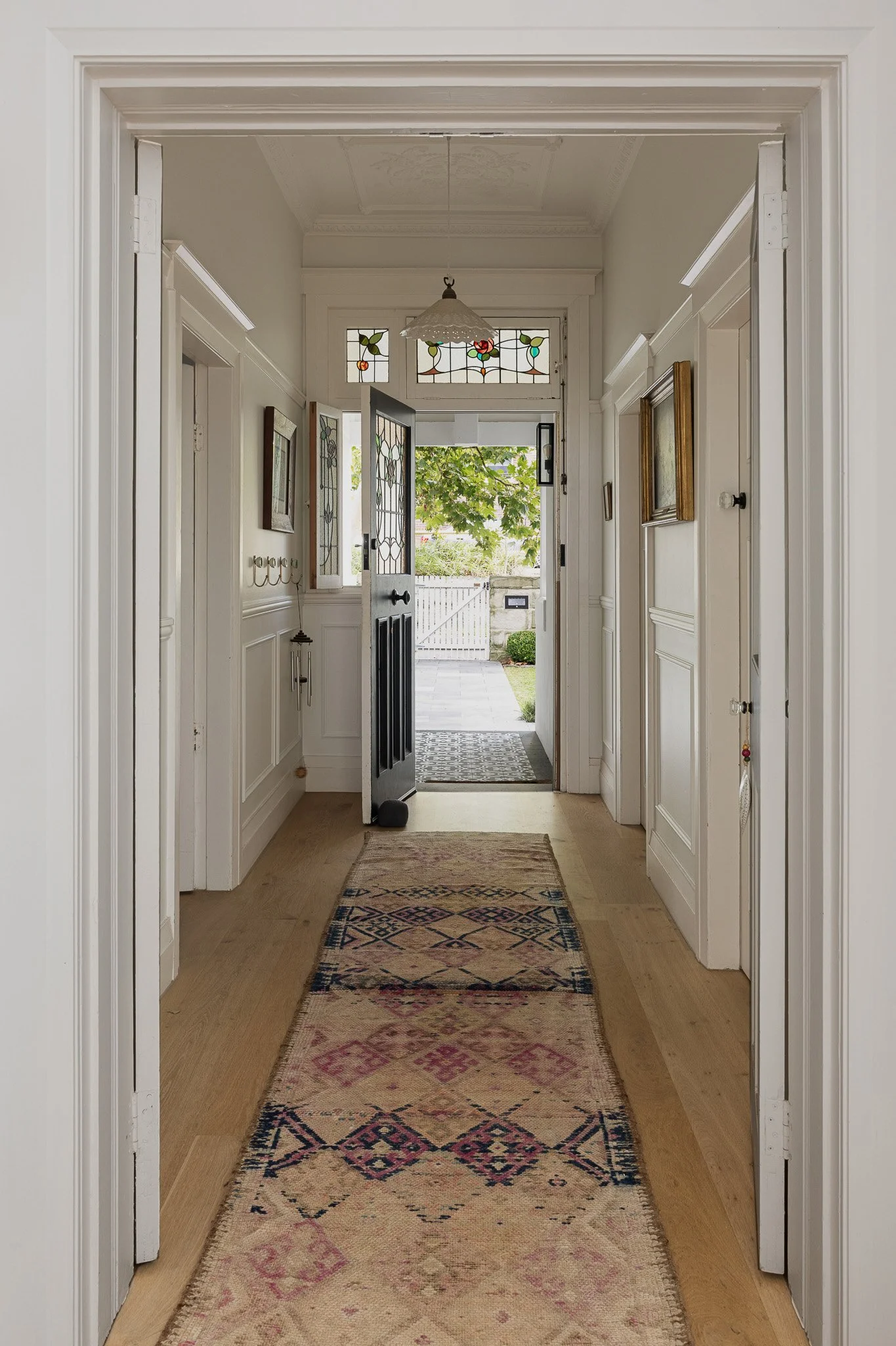 View of a home's hallway with an open front door leading to a yard with a white fence and trees, and a patterned rug on the wooden floor.