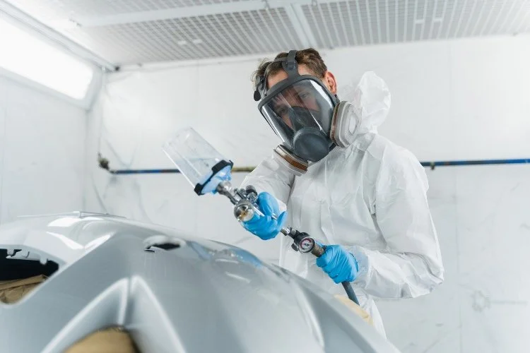 A man in a white suit and mask is painting a car, focused on applying a fresh coat of paint.