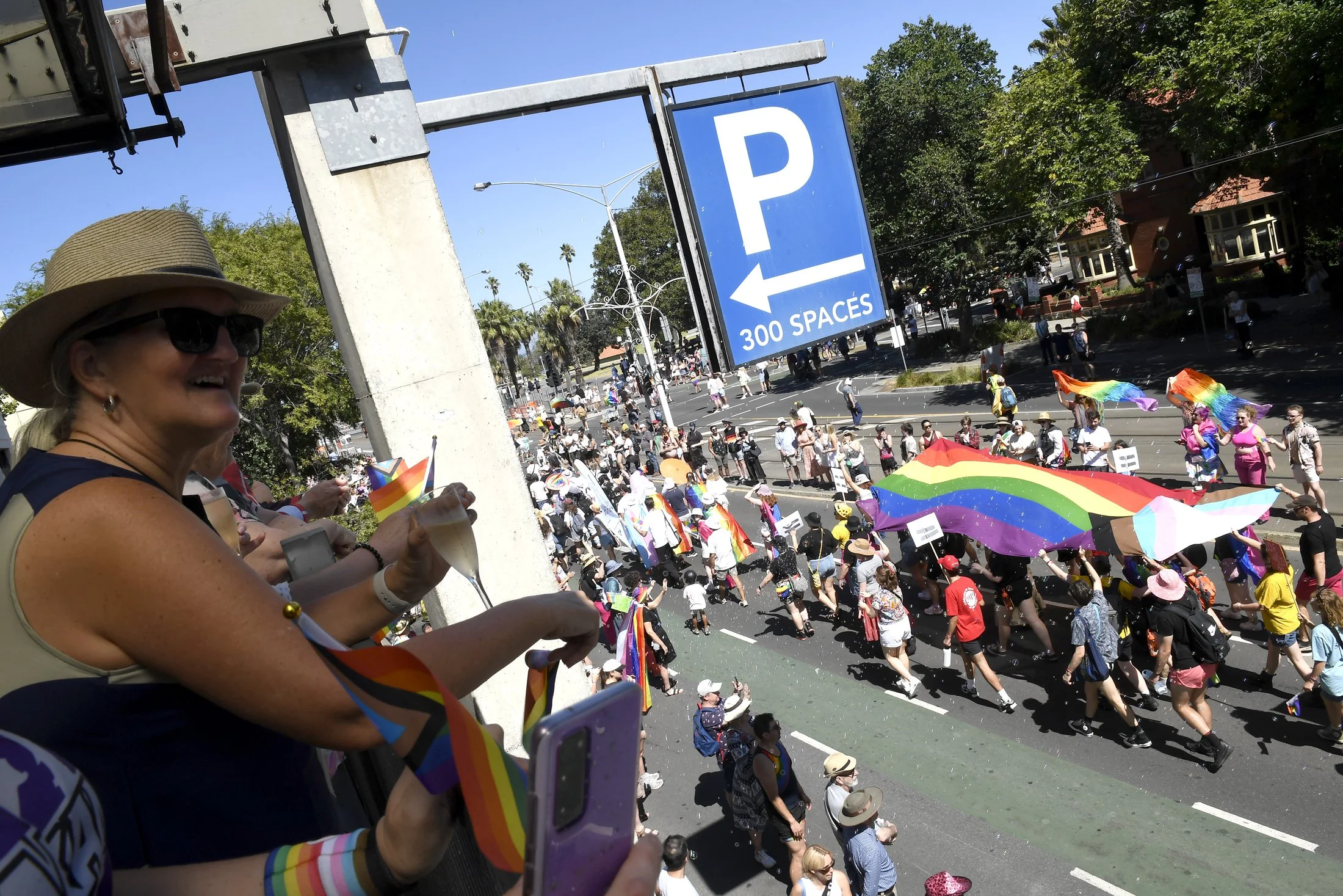 Crowd of people participating in a pride parade, holding rainbow flags and umbrellas, with some on a balcony overlooking the street.