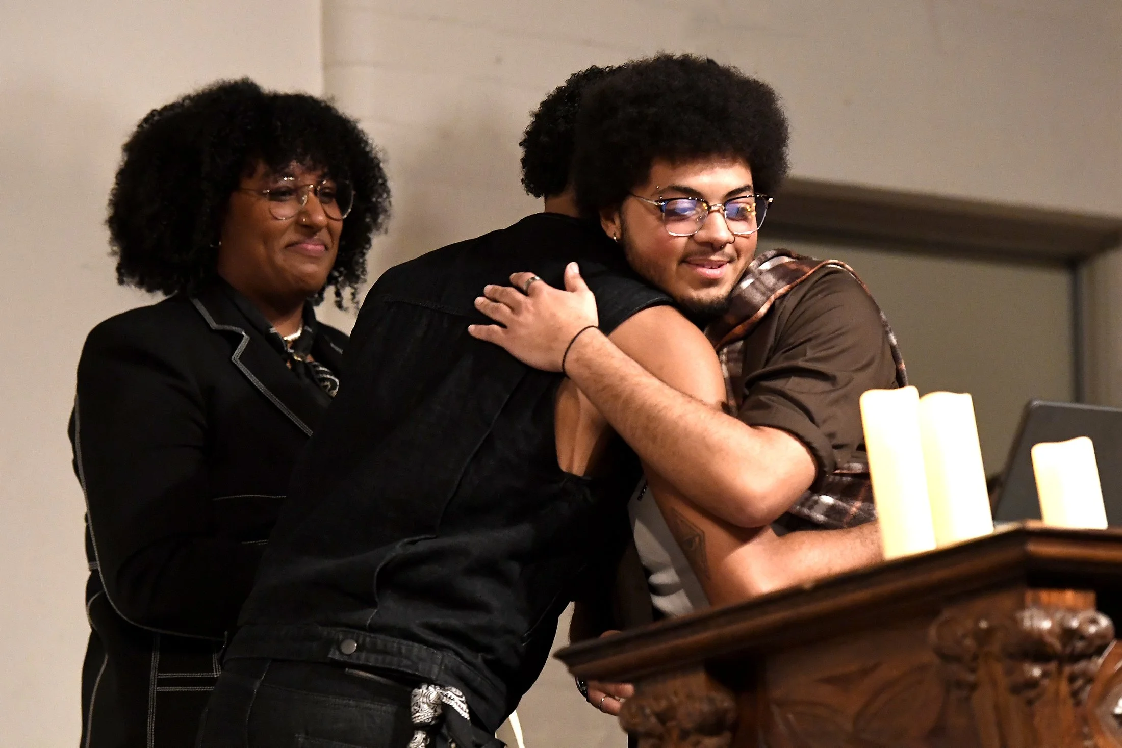 Two men hugging each other while a woman looks on with a smile in an indoor setting with candles on a wooden surface.