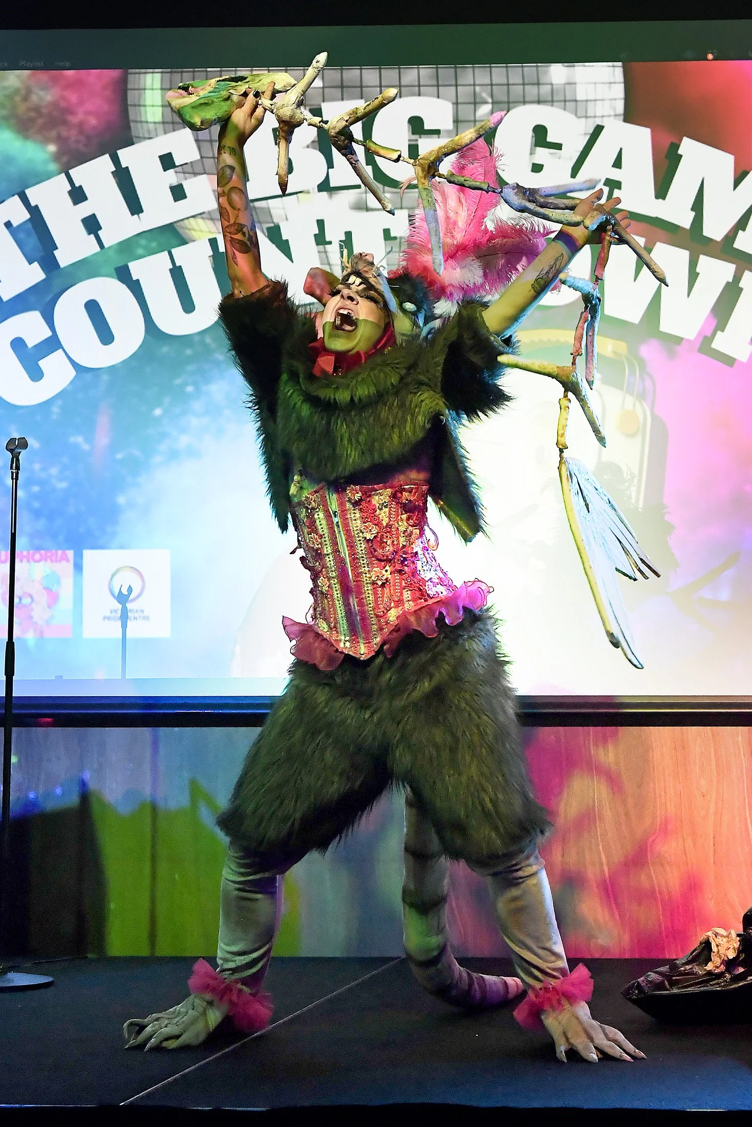 Performer dressed as a fantastical creature with animal-like features, holding a branch with colorful elements during a stage performance.