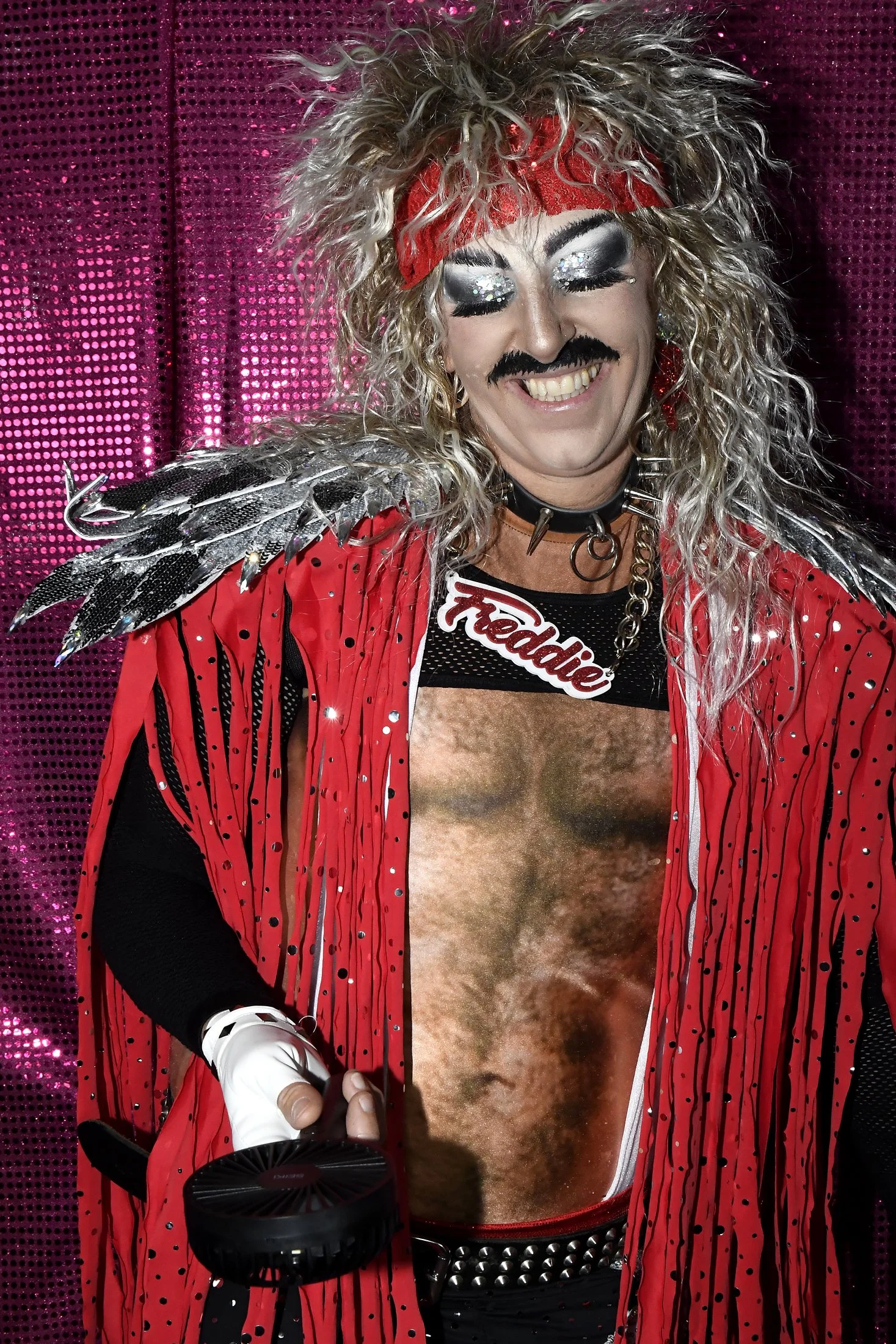 A person dressed in a colorful, rock-inspired costume with extravagant makeup, a red headband, and a name tag that says 'Feddie.' They are smiling with eyes closed against a pink, sequined backdrop and holding a small fan.
