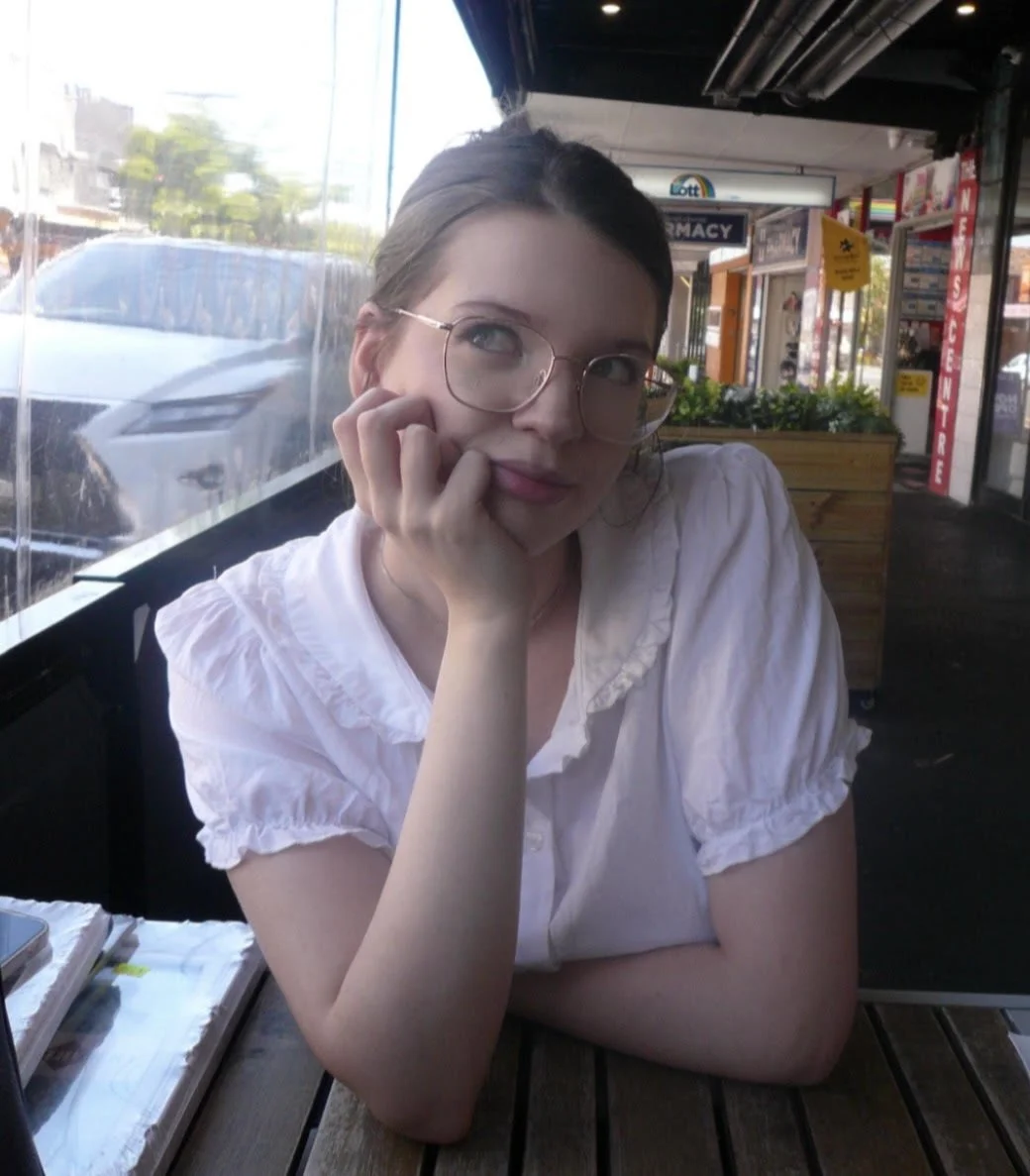 A young woman with glasses, wearing a white blouse, sitting at a table in a cafe or restaurant, with her face resting on her hand and looking thoughtfully outside.