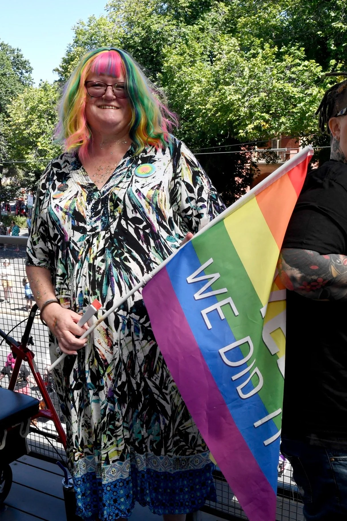 Person with rainbow-colored hair holding a rainbow pride flag at an outdoor LGBTQ+ event.