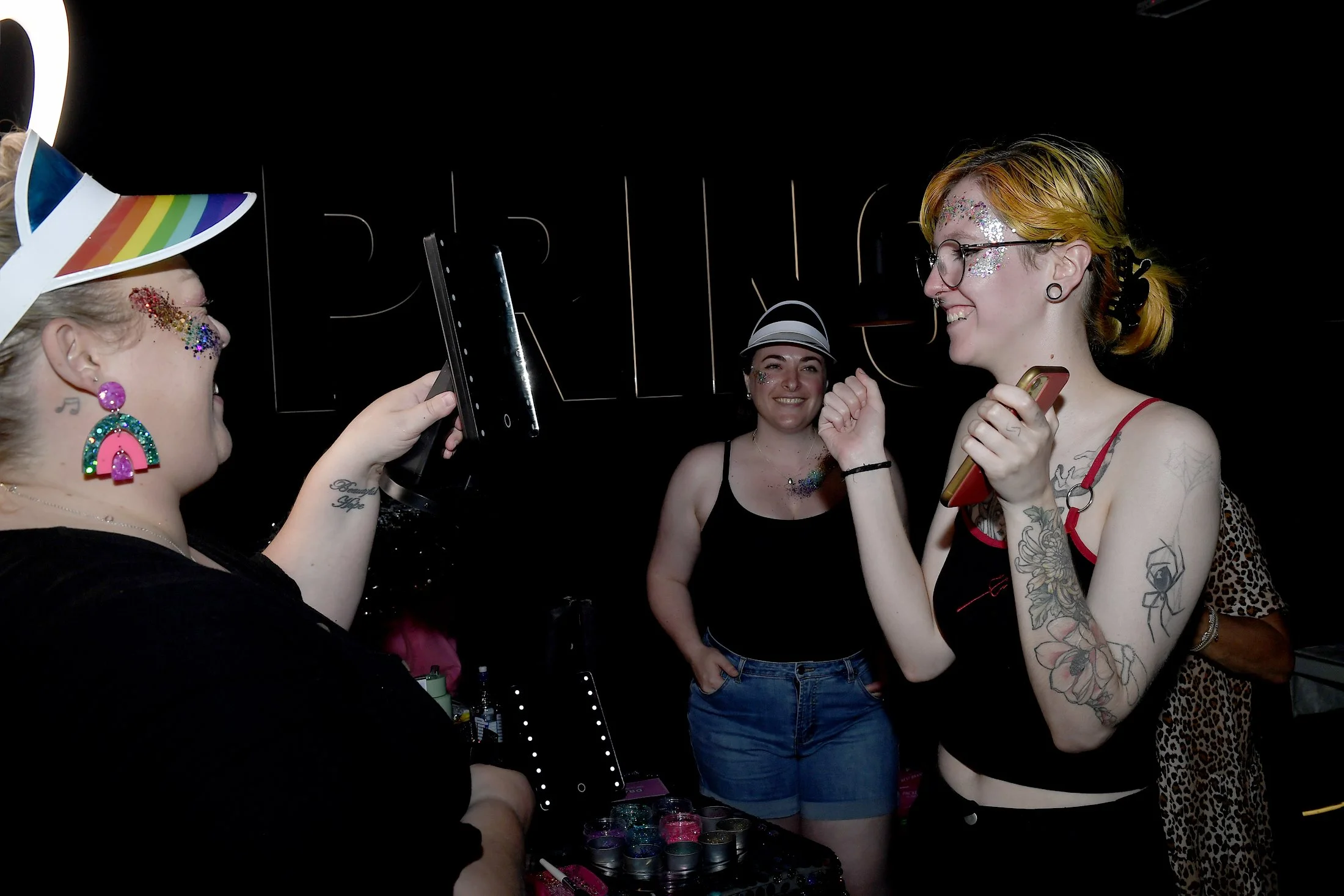 Three women at a party, one wearing a rainbow hat and colorful earrings, another with glitter on her face and glasses, and the third in a black tank top and shorts, smiling and holding a phone, in front of a black background with neon light "BRIDGE."