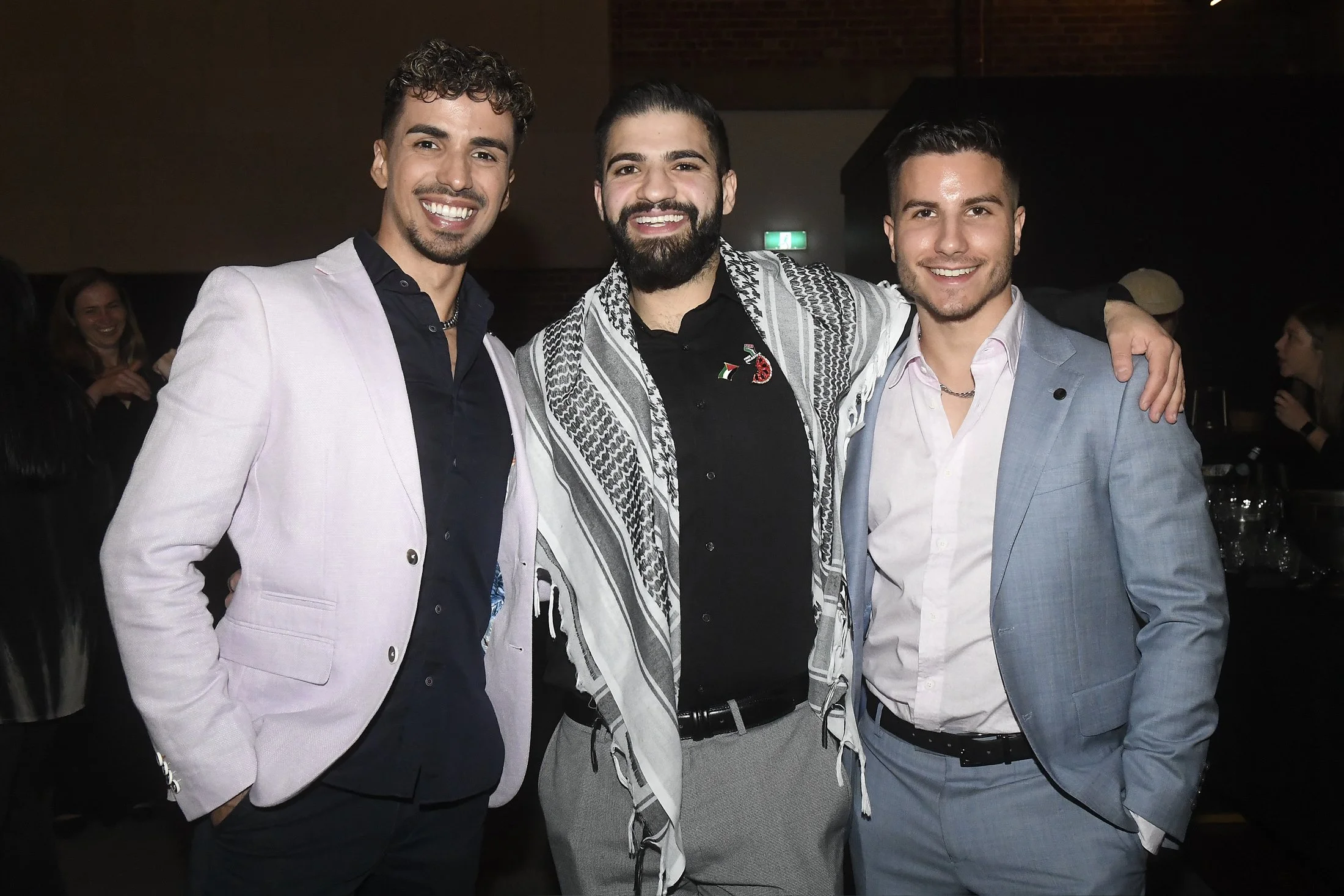 Three men in suits smiling with arms around each other at an indoor event