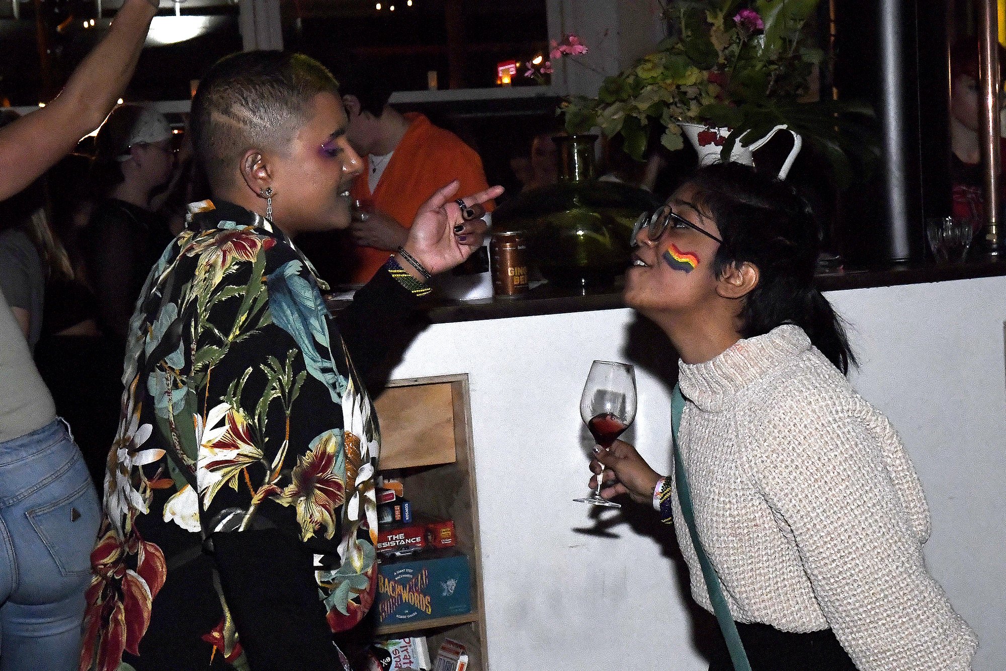 Two people at a bar or party, one with rainbow face paint and holding a glass of red wine, and the other with multicolored makeup, in conversation.