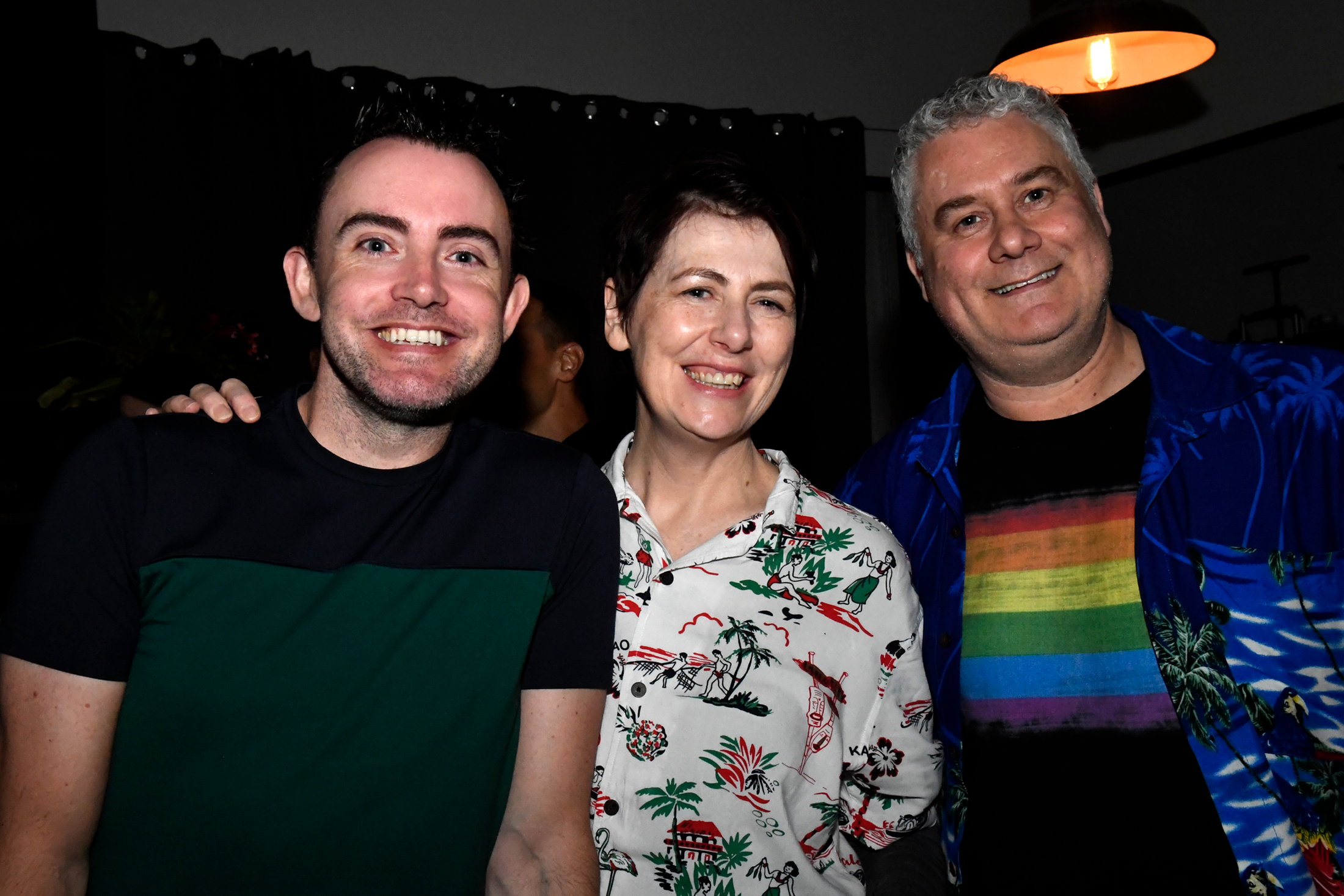 Three smiling people standing together at a social gathering, with a black curtain and warm lighting in the background.
