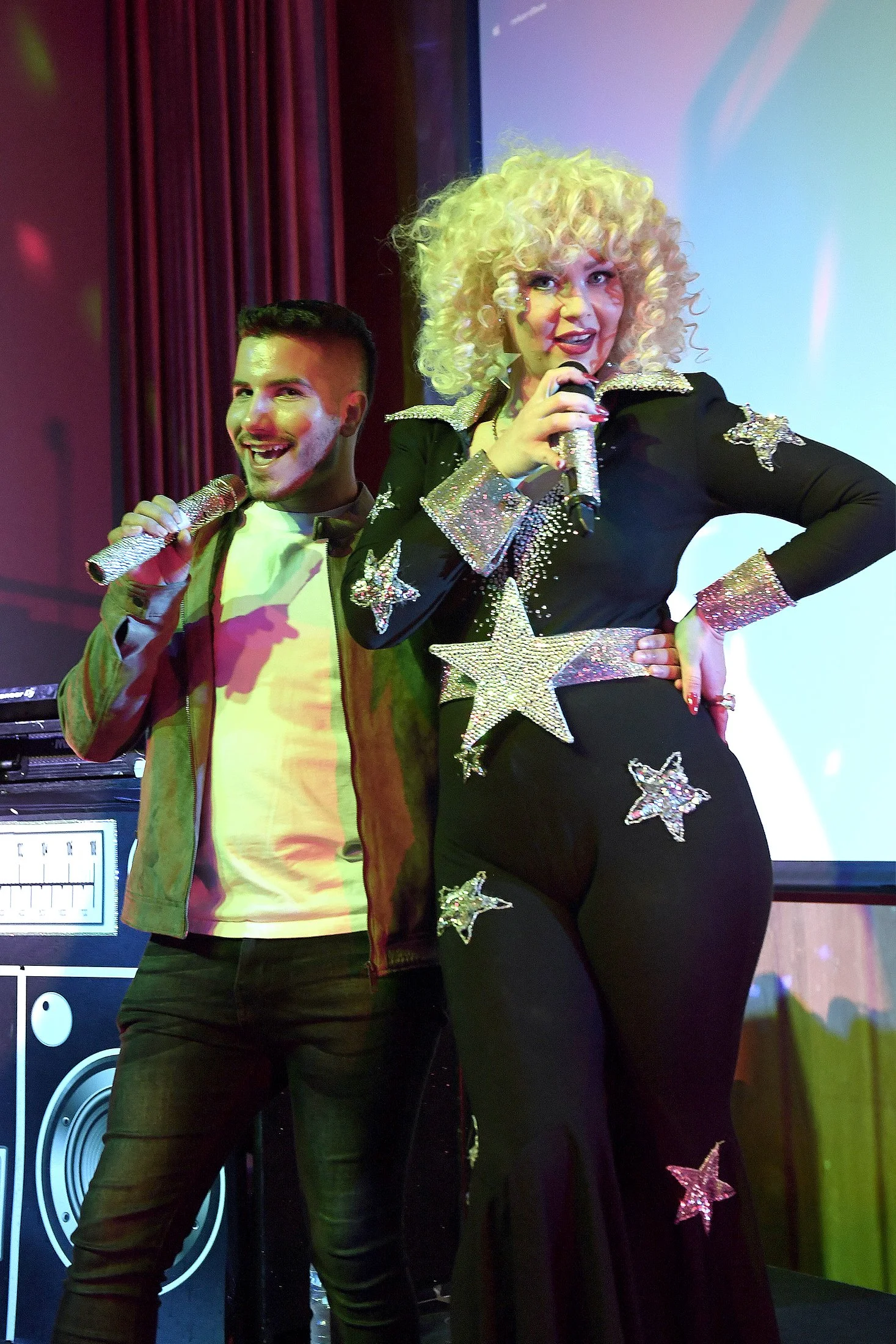 Two performers on stage, a man and a woman, holding glittery microphones. The woman has curly blonde hair, is dressed in a black outfit with large star-shaped embellishments, and is speaking into the microphone. The man has short dark hair, is wearin
