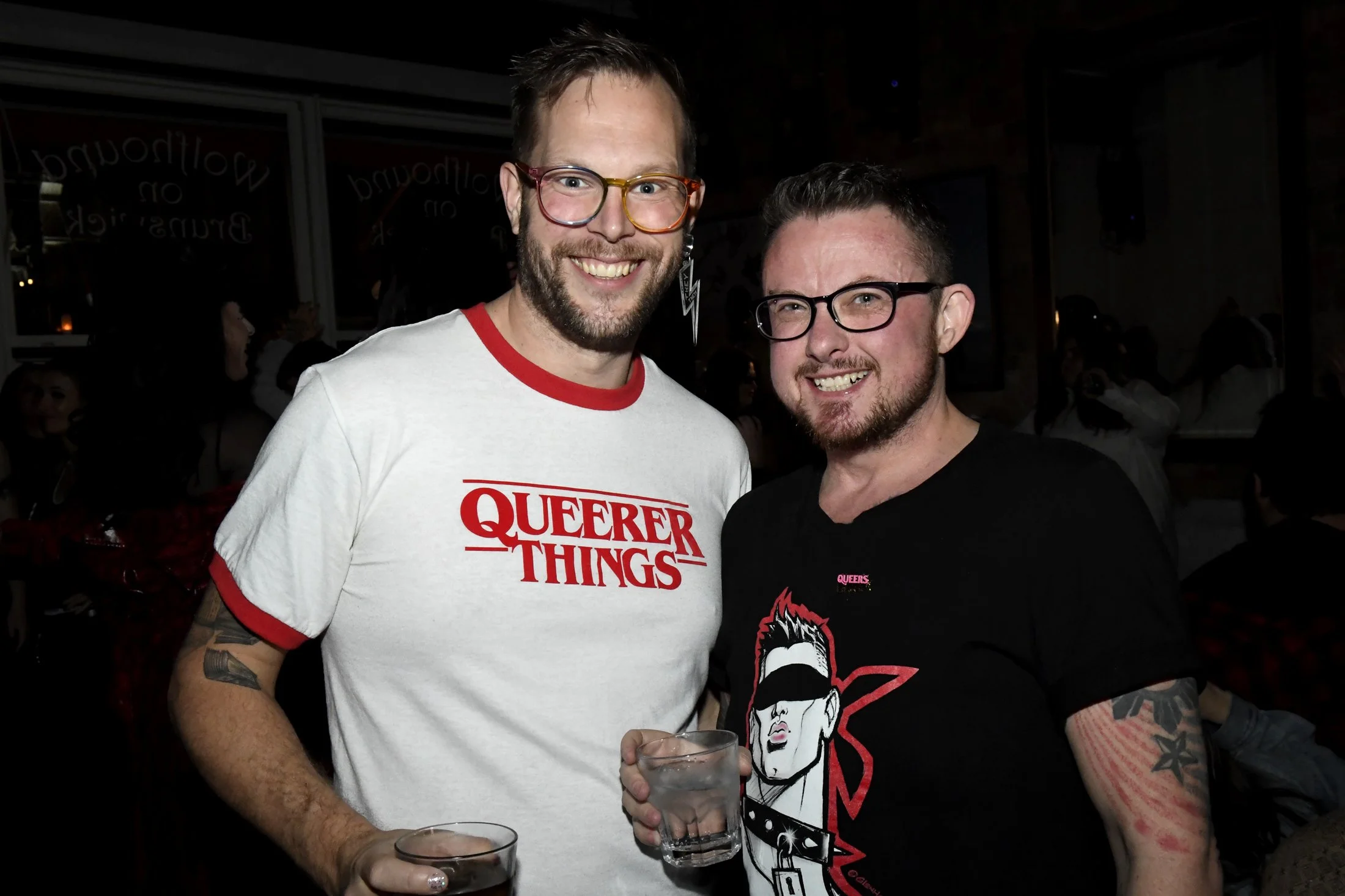 Two men smiling and standing together at a party, each holding a drink, with a dark background and other people in the distance.
