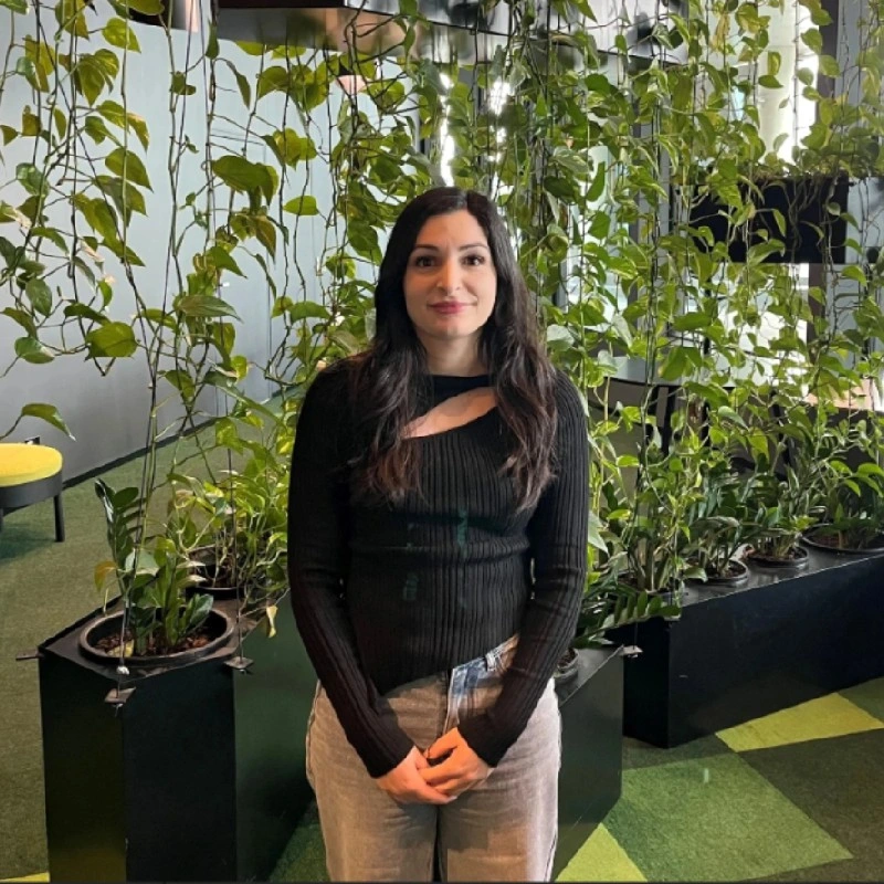 A woman with long dark hair wearing a black long-sleeve top and beige pants standing in front of green-leafed plants in a modern indoor space.