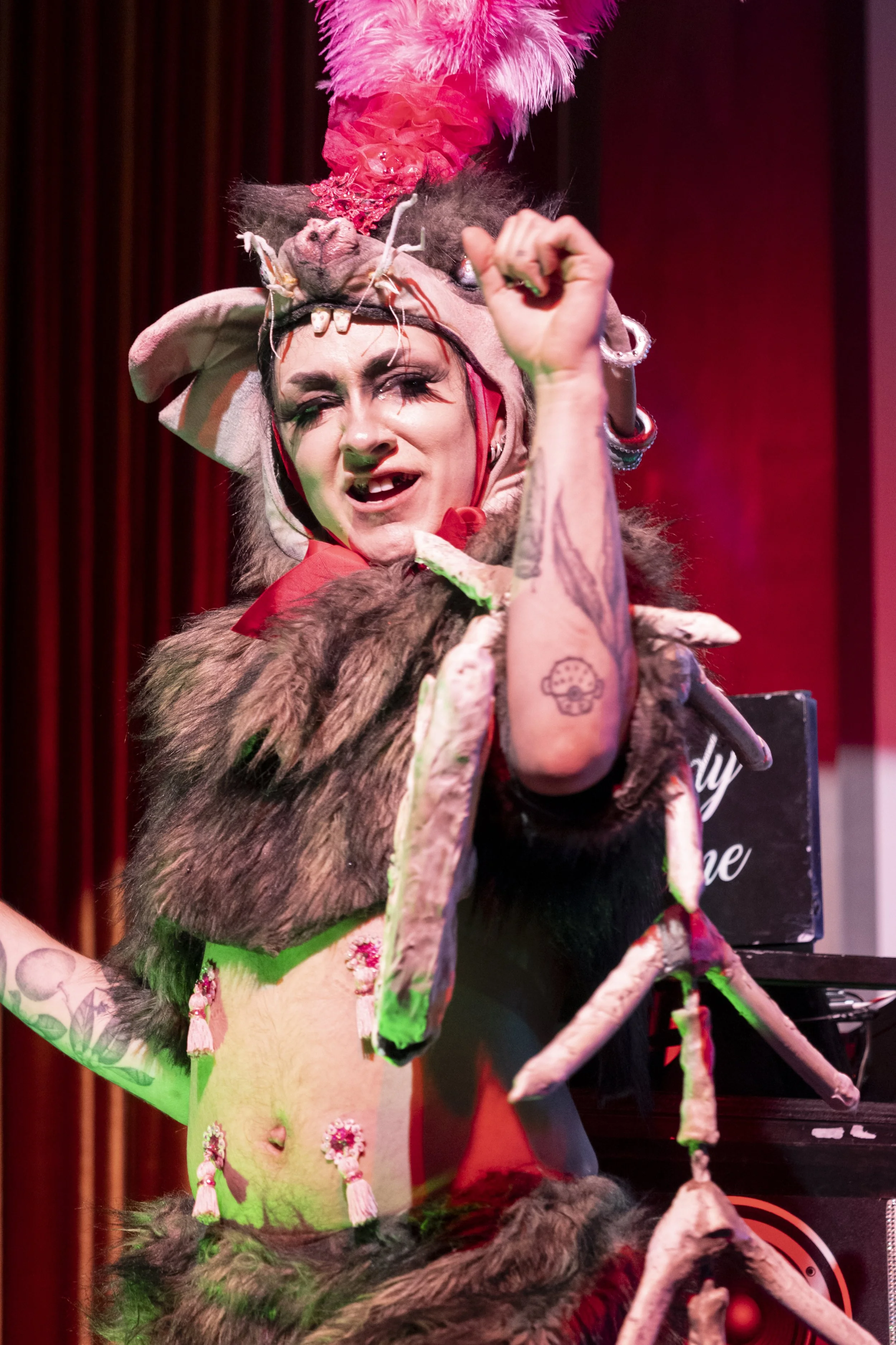 Performer in elaborate costume with animal elements, horns, and feathers, raising fist on stage with red curtain background.
