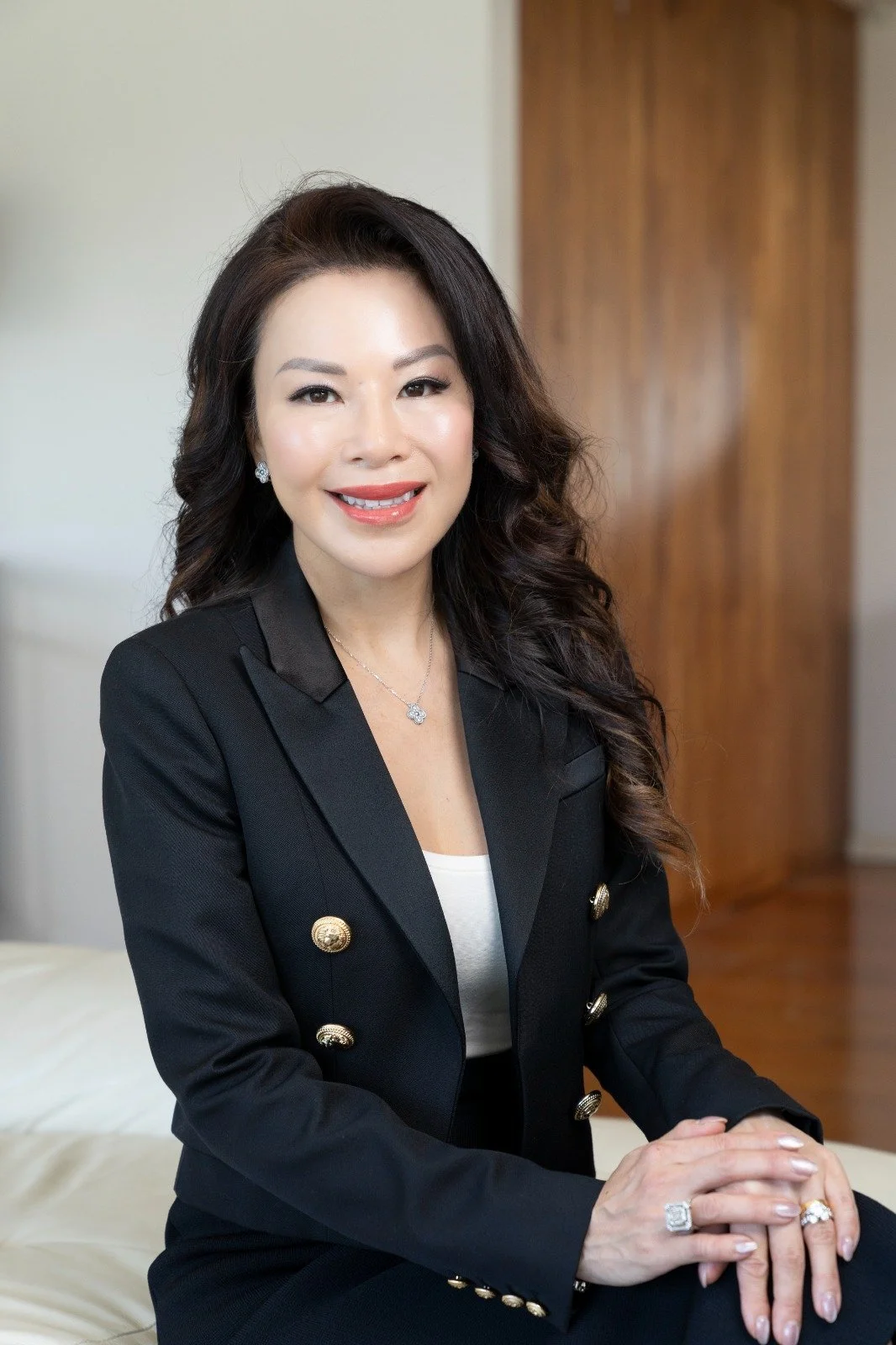 A woman with long dark curly hair, wearing a black blazer with gold buttons, sitting indoors in front of a wooden wall. She is smiling and wearing jewelry including earrings, a necklace, and rings.