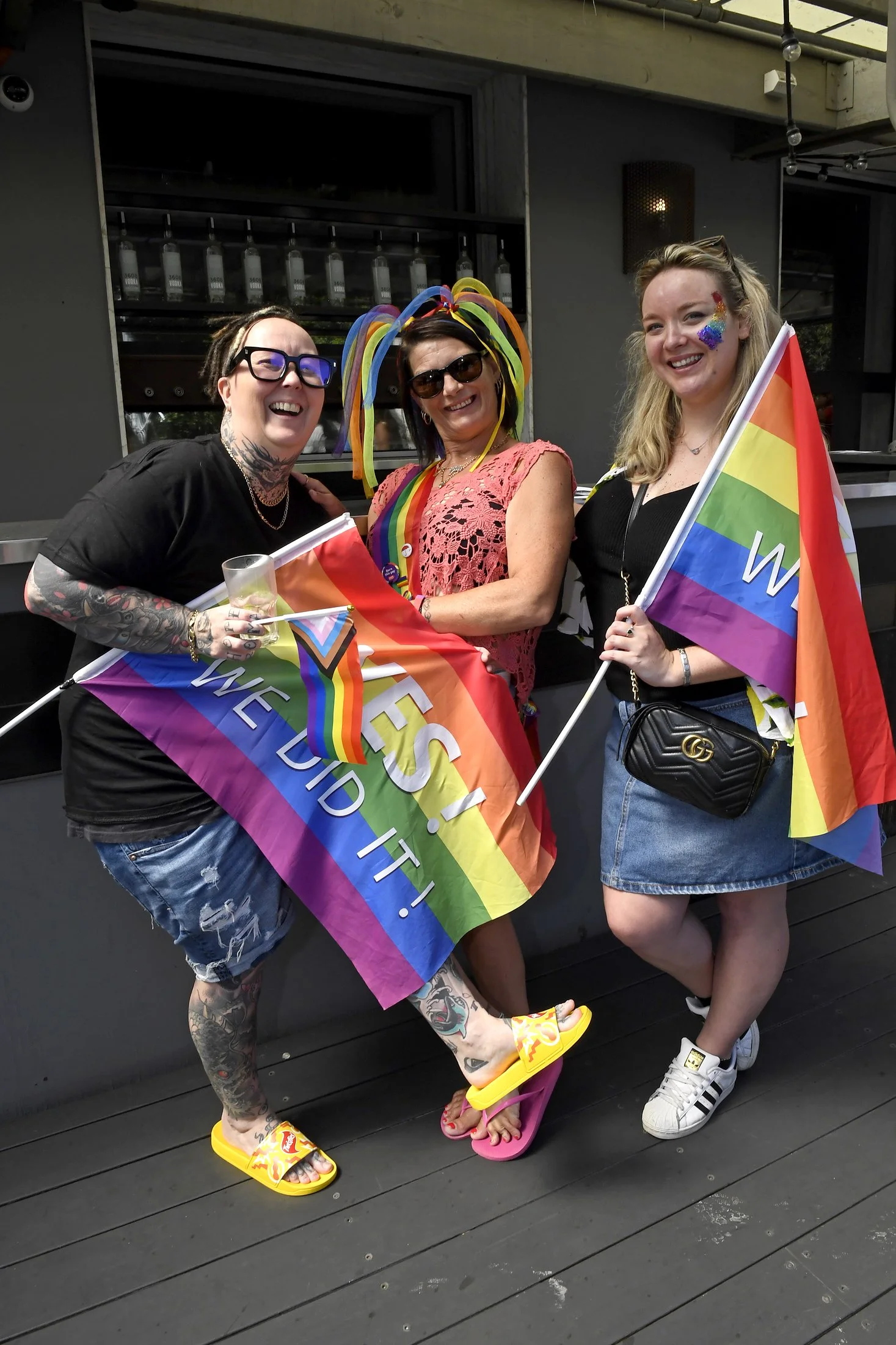 Three women celebrating LGBTQ pride outdoors, holding rainbow flags, wearing pride-themed accessories and clothing, smiling and enjoying the event.