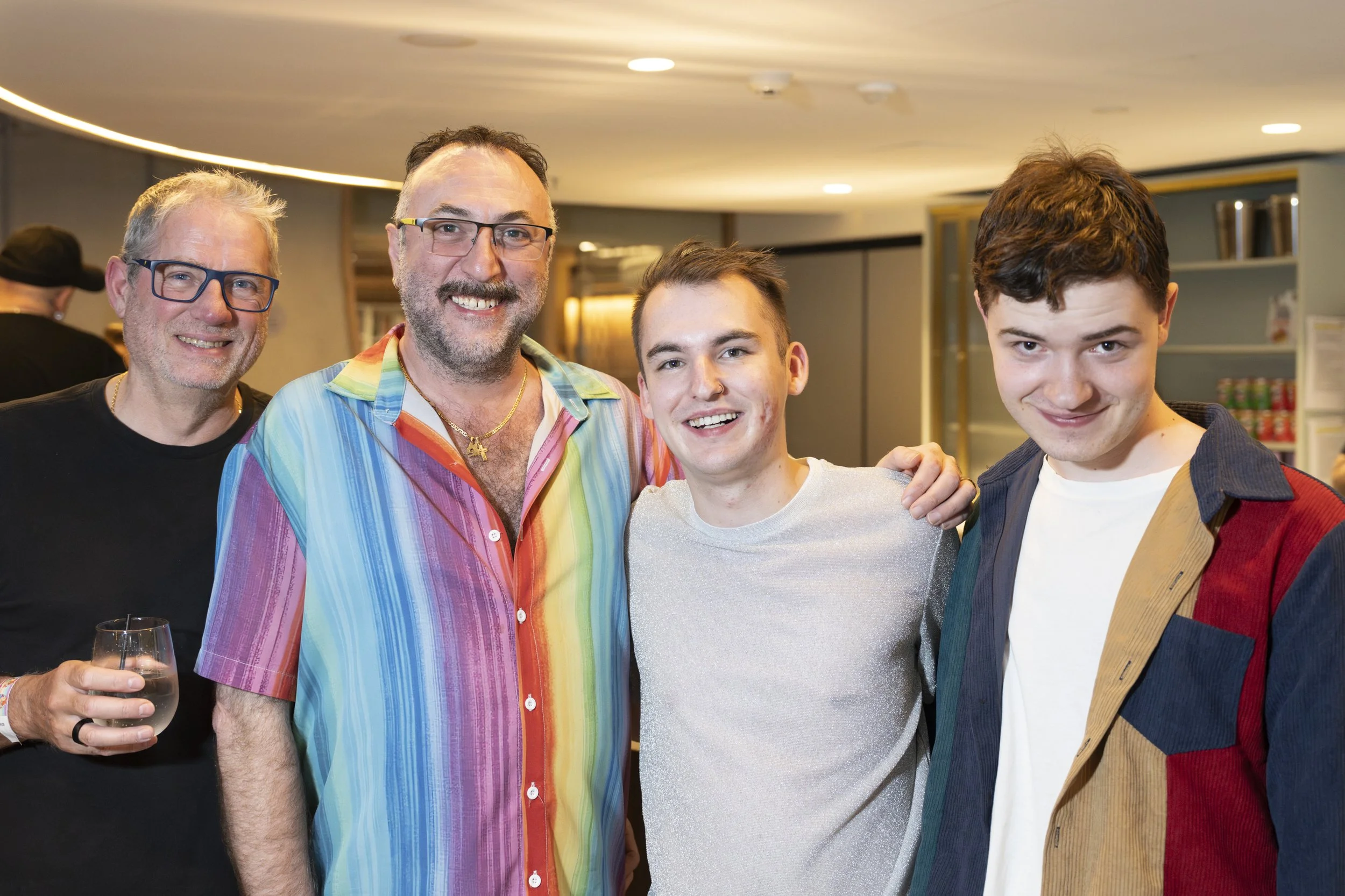 Four men smiling at a gathering, standing close together indoors, with bookshelves and beverages in the background.