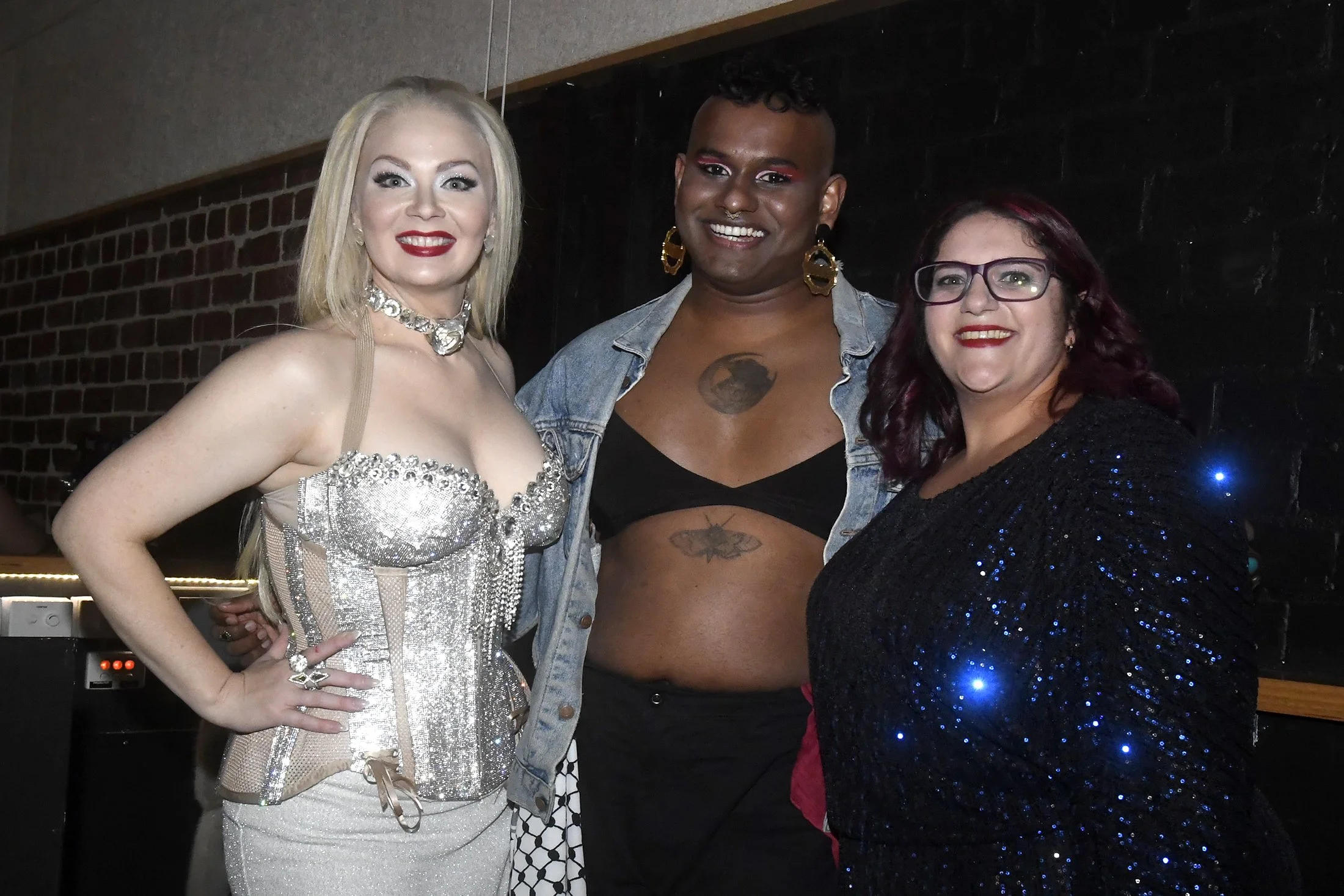 Three women posing together indoors, dressed in glamorous and colorful outfits with accessories, smiling at the camera.