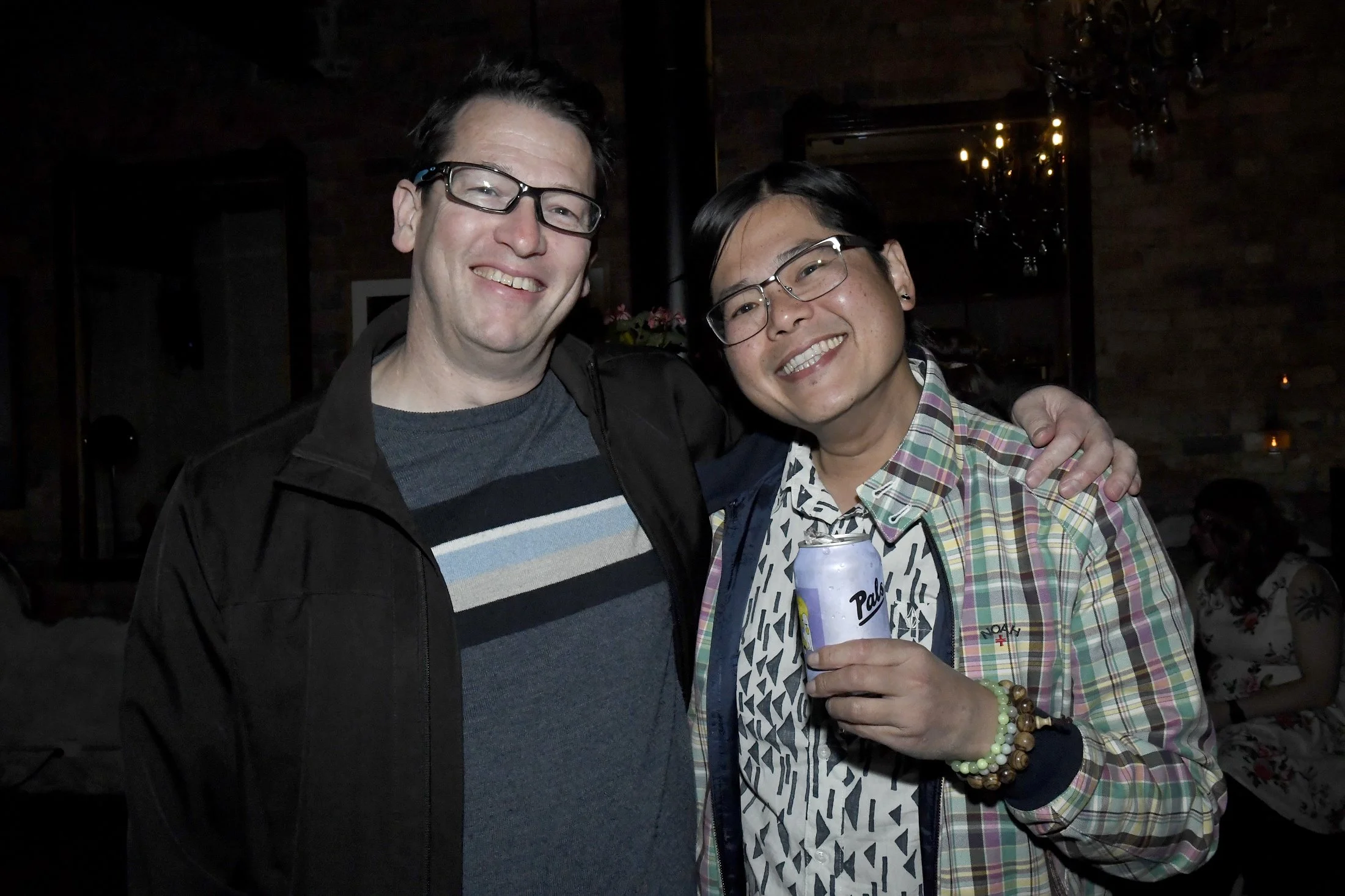 Two men smiling at a social gathering, one with short dark hair and glasses, wearing a dark jacket and striped shirt, and the other with short black hair and glasses, wearing a plaid shirt with a patterned hoodie, holding a can of Pabst Blue Ribbon b