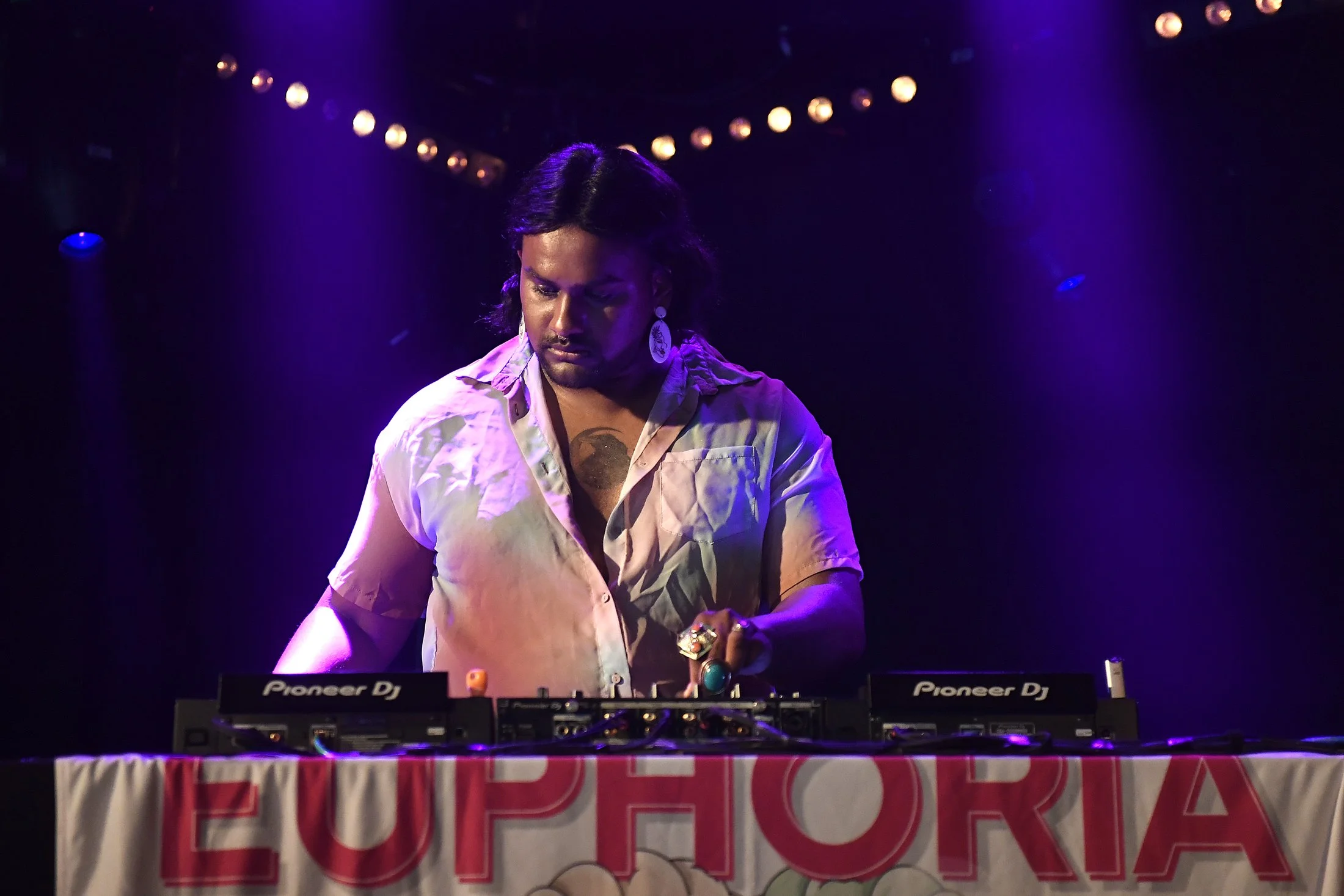 A DJ performing at a music event, wearing rings and earrings, with purple stage lighting and a banner that reads 'Euphorica'.