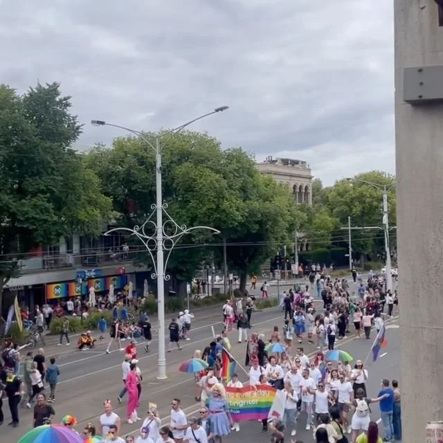 A street scene with a large crowd of people participating in a parade, holding rainbow flags and banners, with trees, buildings, and overcast sky in the background.