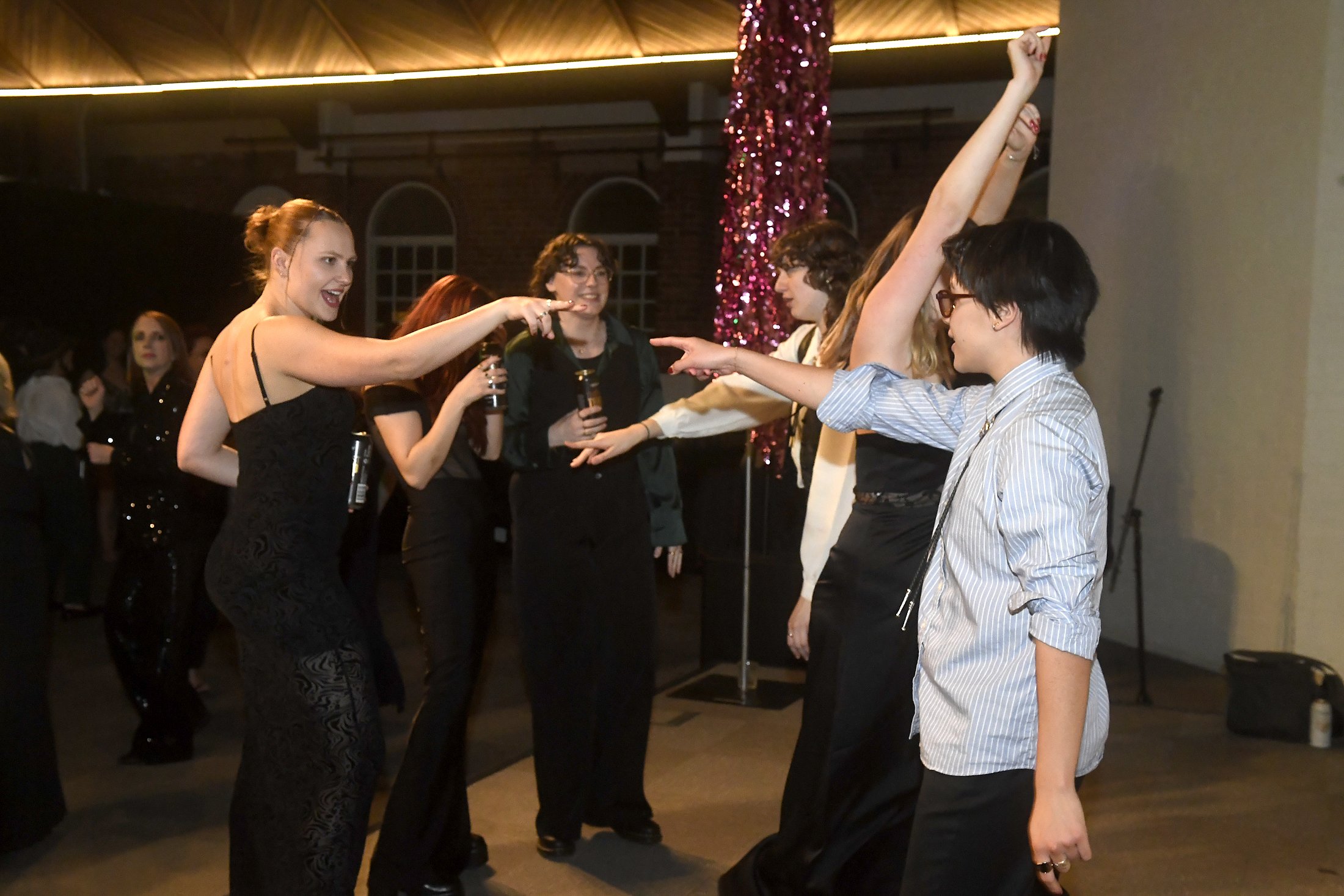 People dancing and enjoying themselves at a party, with some holding drinks, in a festive indoor setting with pink decorations.